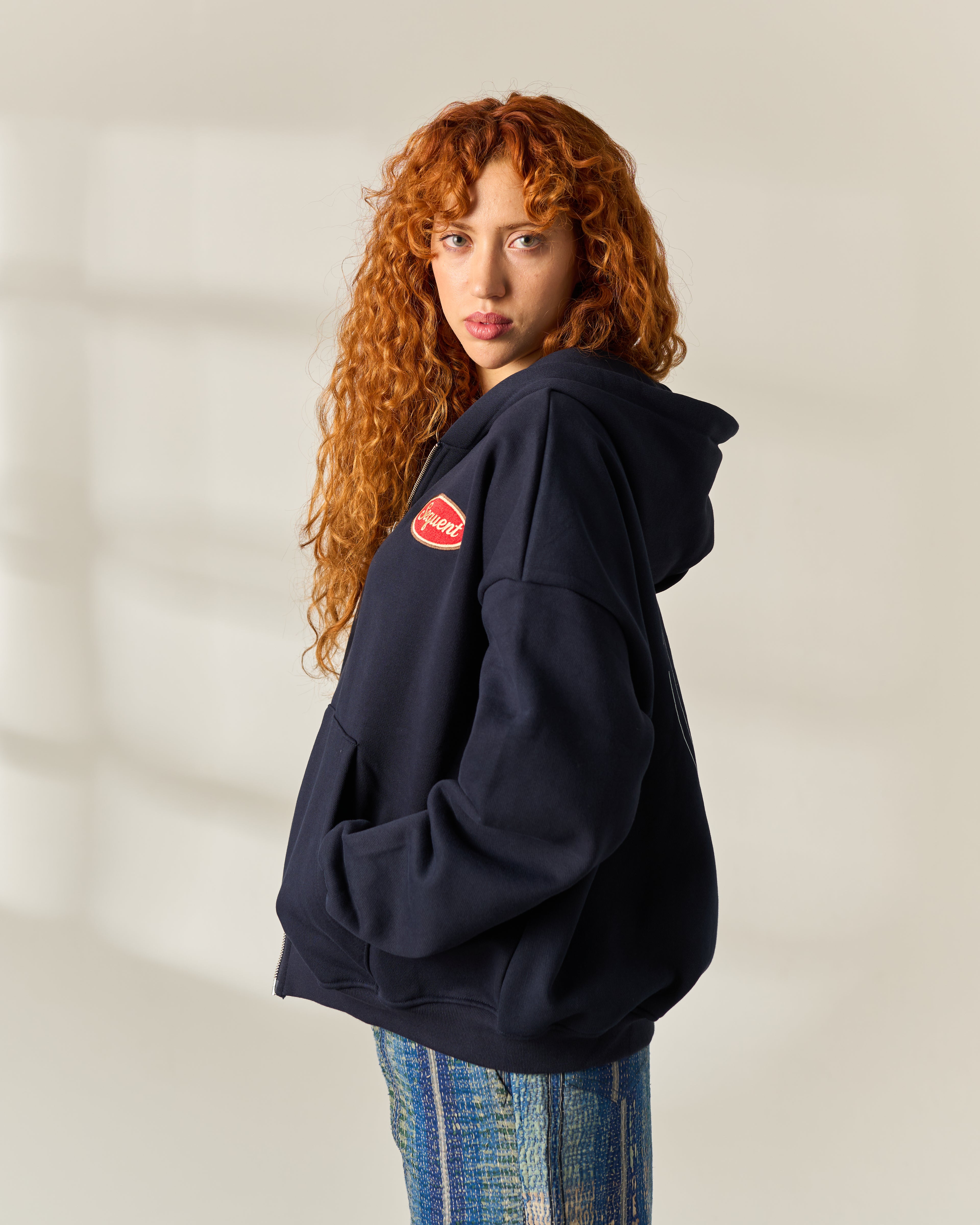 A woman with long curly red hair is shown from the back wearing an oversized navy blue hoodie with a red circular patch on the left shoulder, paired with blue jeans, against a warm off-white background with soft window shadows.