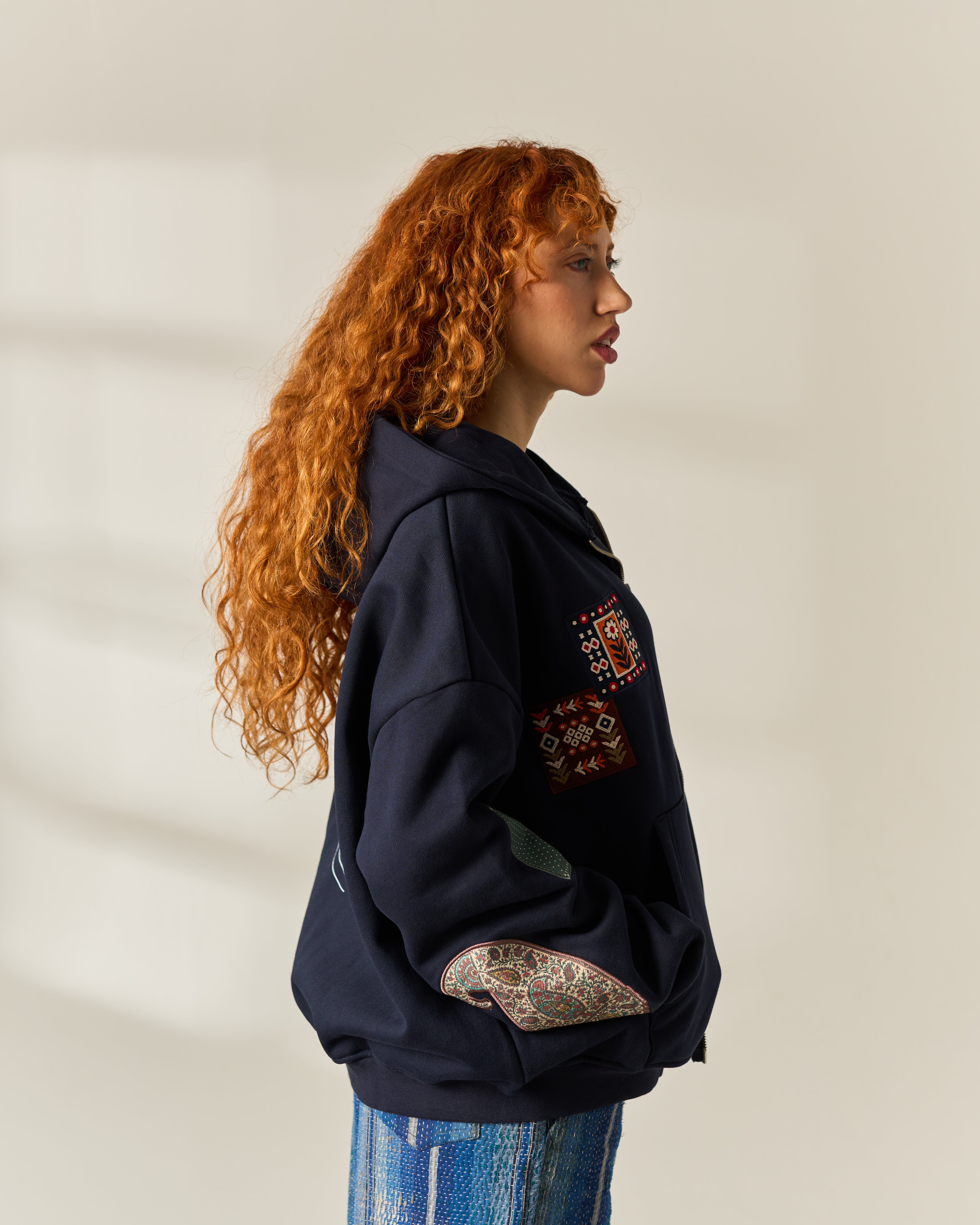 A woman with long, curly red hair is shown in profile wearing a navy blue hoodie with colorful embroidered patches on the chest and sleeve, and patterned knit elbow patches. 