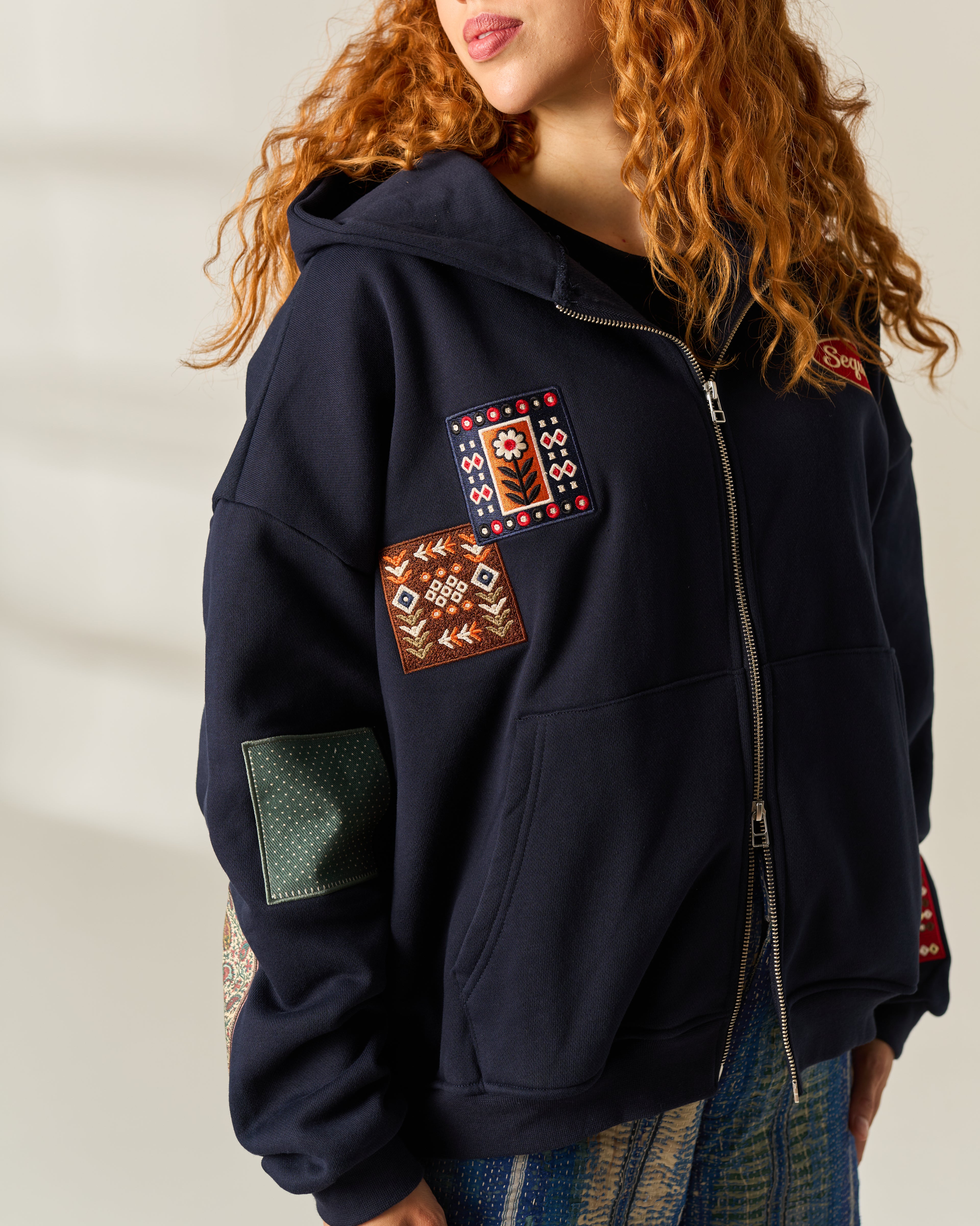 A close-up of a person wearing a navy zip-up hoodie with multiple colorful embroidered and patch details on the front and sleeves, paired with patterned blue jeans against a softly lit neutral background.