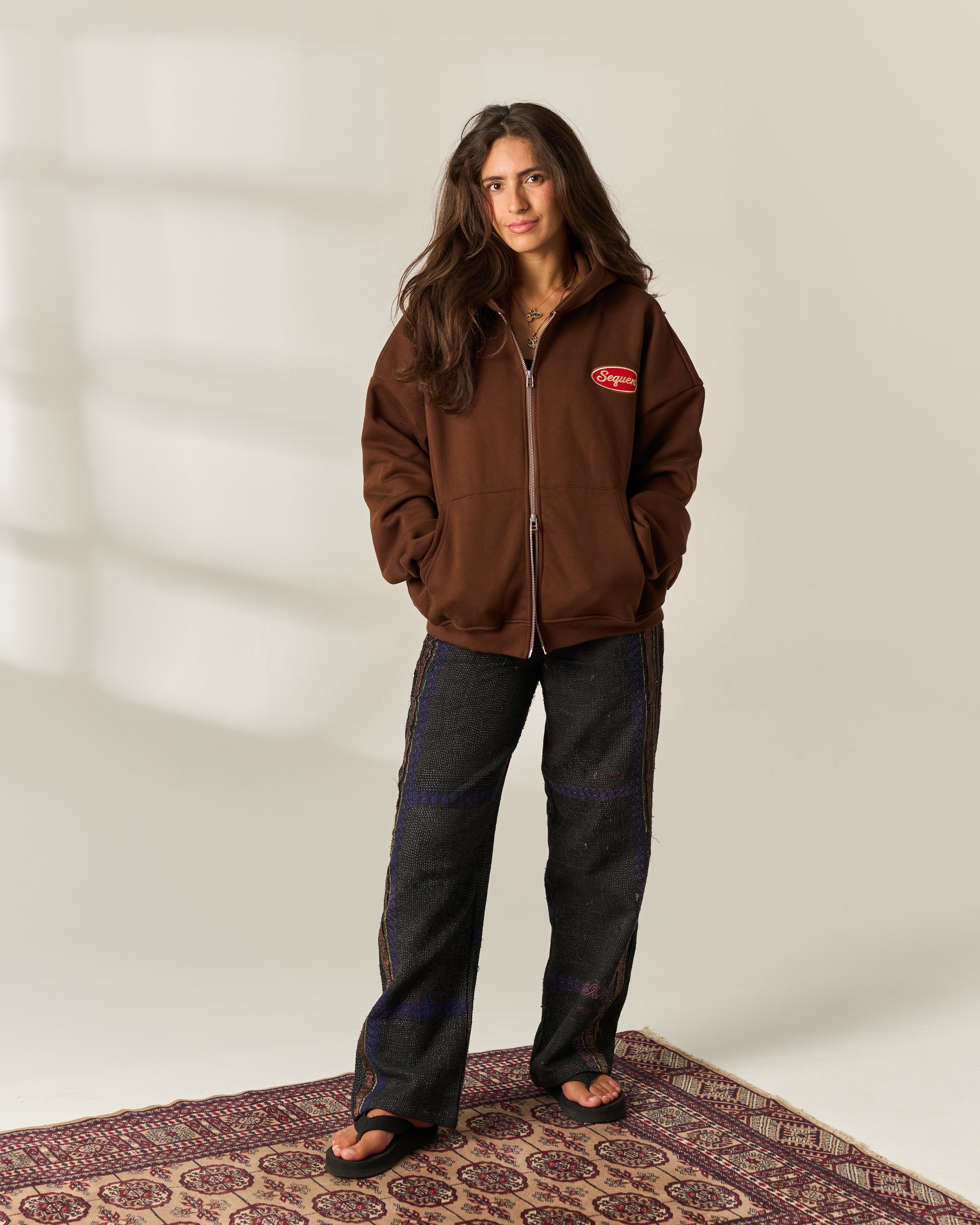 A woman stands indoors wearing a brown zip-up fleece jacket with a small red chest 'Sequent' logo, dark wide-leg pants, and black sandals, posed on a patterned rug against a light neutral wall.