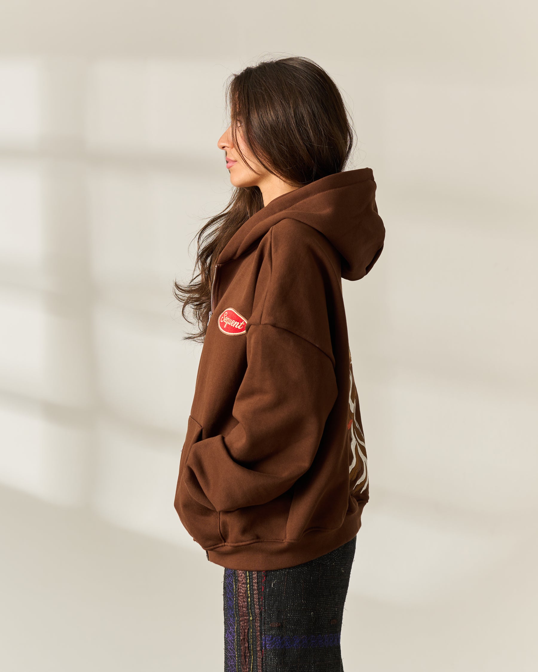 A woman in profile wearing a brown pullover hoodie with a red embroidered patch on the left chest and a graphic embroidered on the back, paired with dark patterned pants against a softly lit beige background.