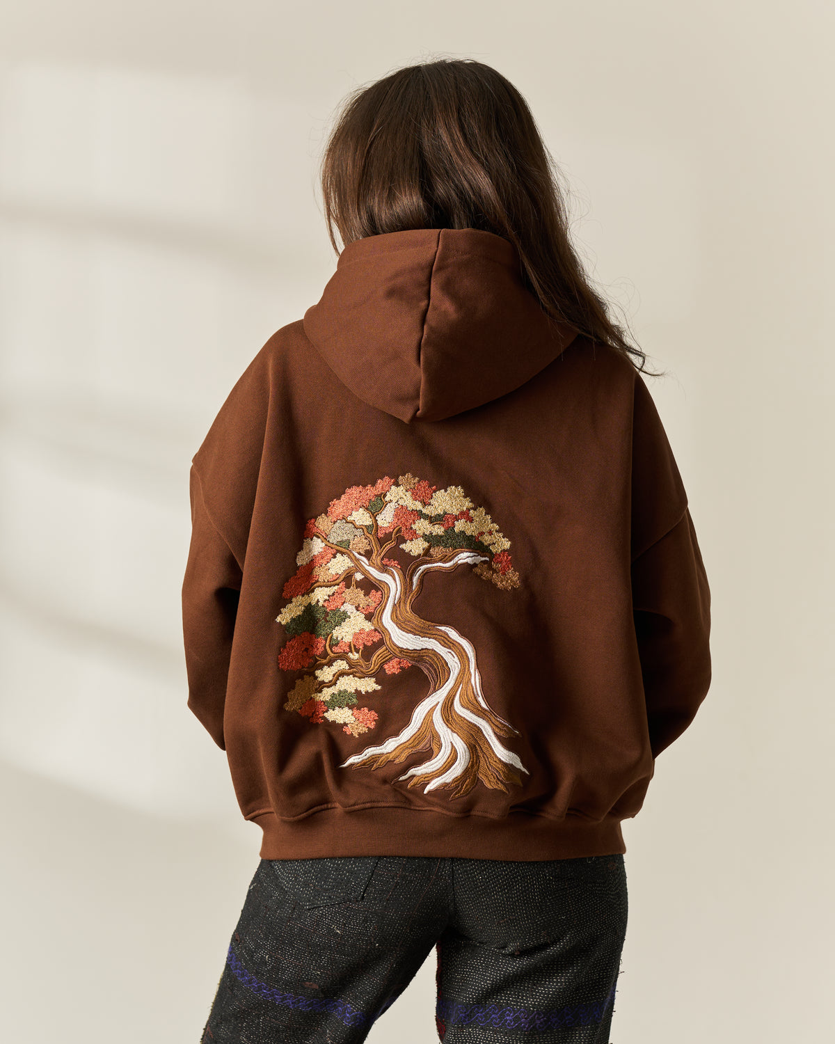 A person viewed from behind wearing a brown hoodie with a large embroidered tree design on the back in warm autumn tones, paired with dark gray trousers against a light beige wall.