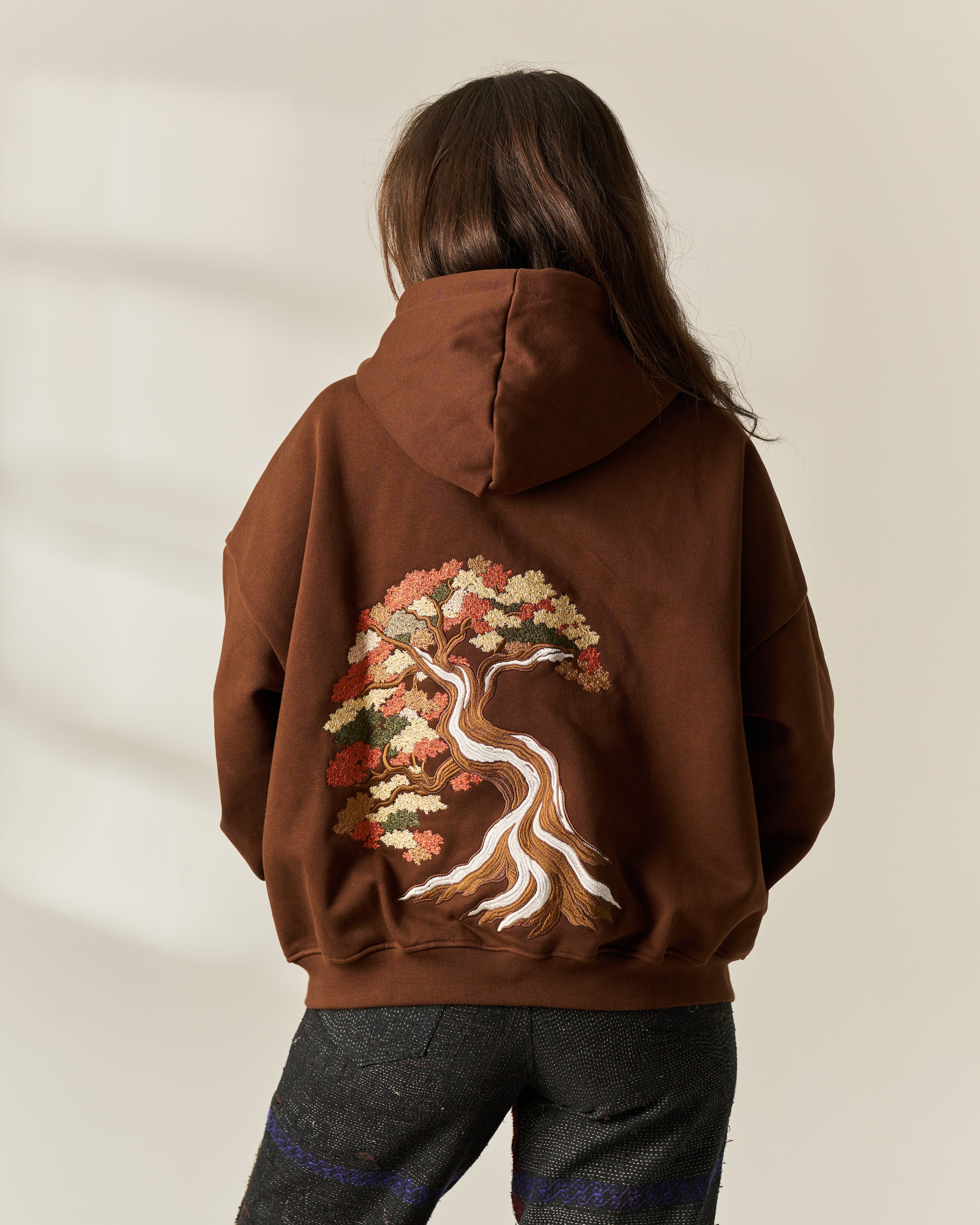 A person viewed from behind wearing a brown hoodie with a large embroidered tree design on the back in warm autumn tones, paired with dark gray trousers against a light beige wall.