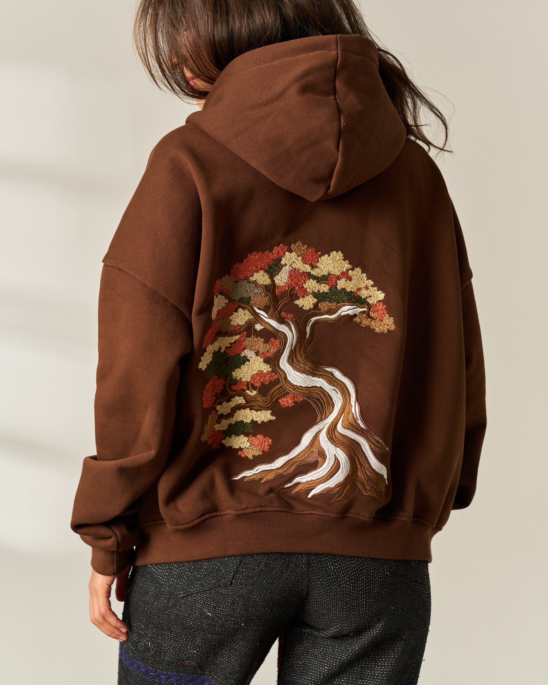 A person shown from the back wearing a brown pullover hoodie with a large embroidered tree design on the back in autumn leaf colors and white, paired with dark gray jeans against a light beige background.