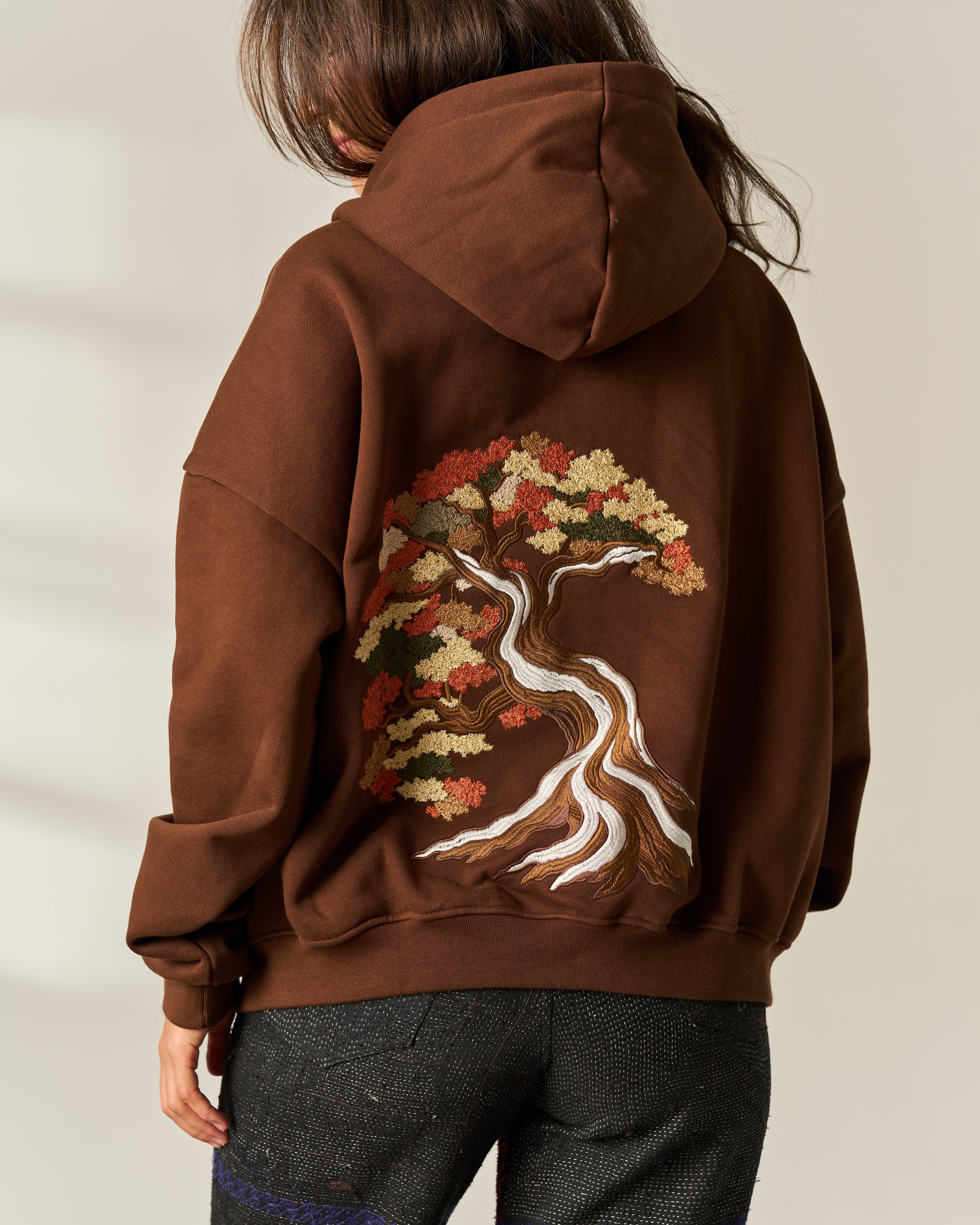A person shown from the back wearing a brown pullover hoodie with a large embroidered tree design on the back in autumn leaf colors and white, paired with dark gray jeans against a light beige background.