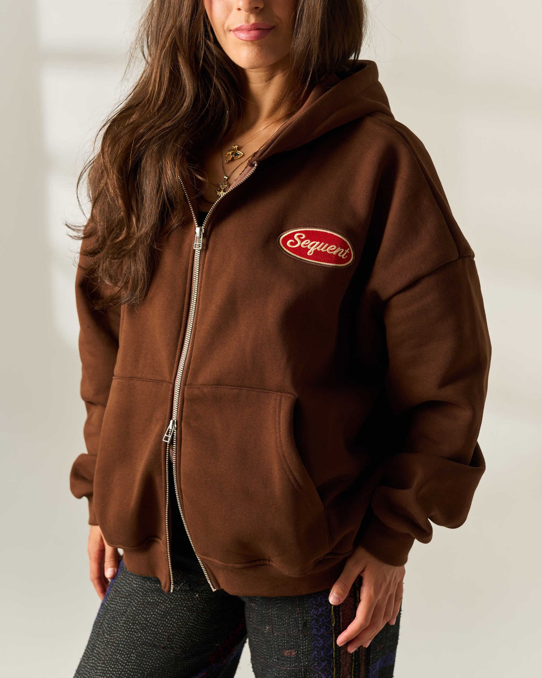 A woman wearing a brown zip-up hoodie with a red oval 'Sequent' embroidered patch on the chest, paired with dark trousers against a neutral indoor background.