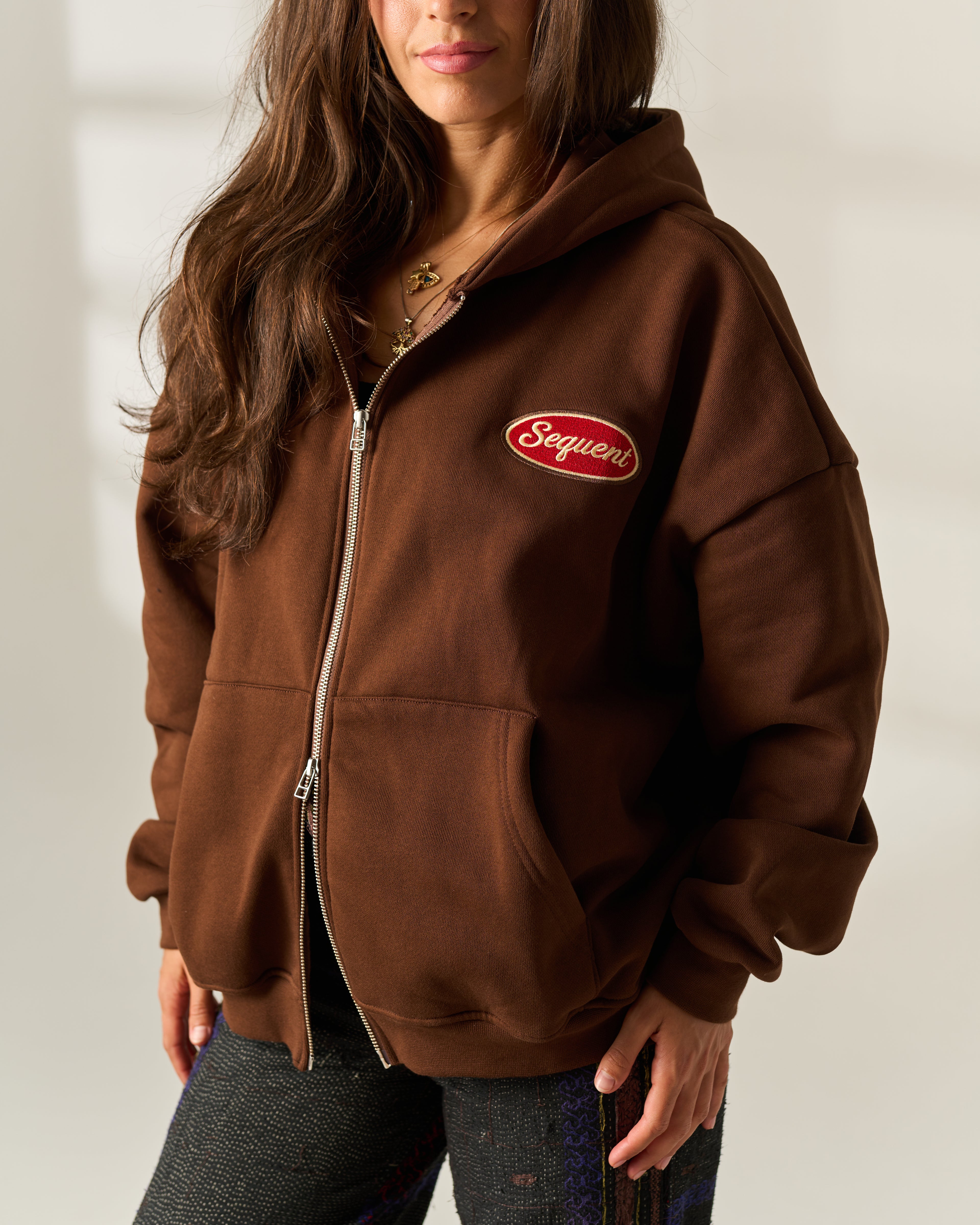 A woman wearing a brown zip-up hoodie with a red oval 'Sequent' embroidered patch on the chest, paired with dark trousers against a neutral indoor background.