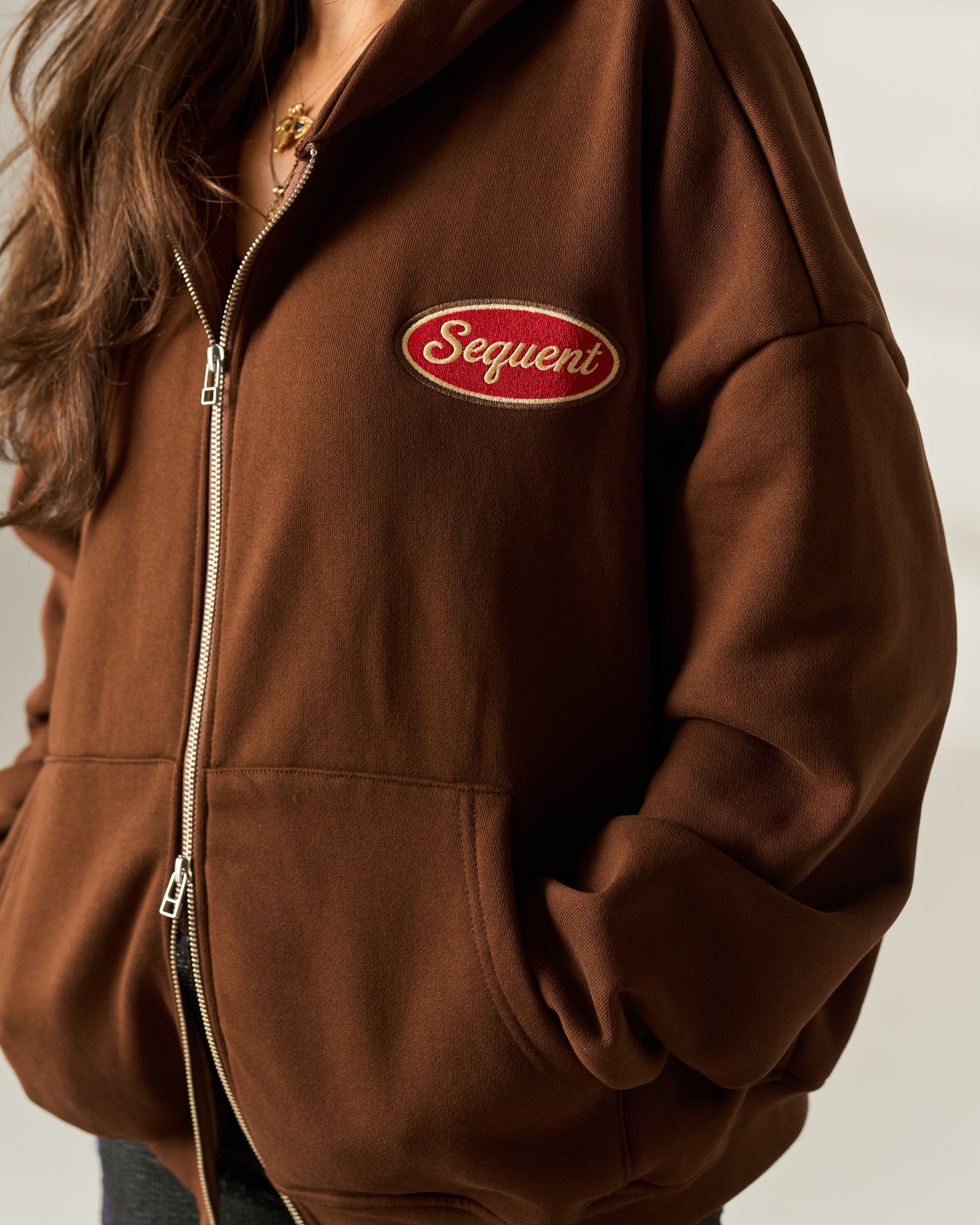 A close-up of a person wearing a brown zip-up hoodie with a red oval 'Sequent' logo patch on the chest, photographed against a plain light background.