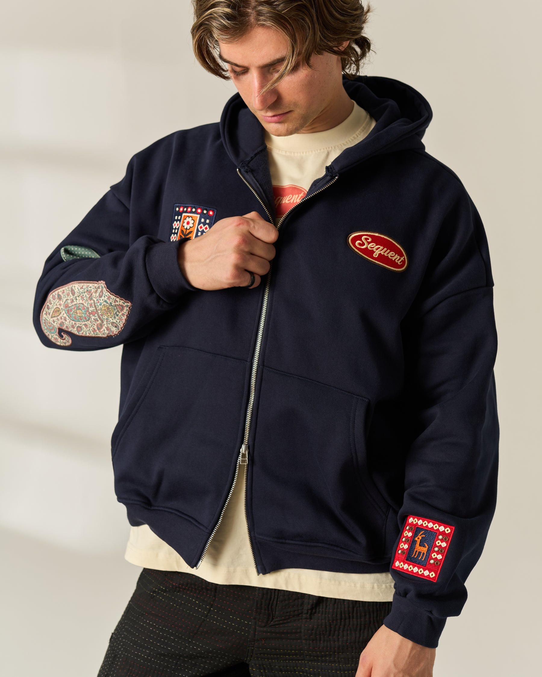 A man wearing a navy blue zip-up hoodie with multiple colorful embroidered patches and a red 'Sequent' logo on the chest, layered over a cream t-shirt and dark pants.