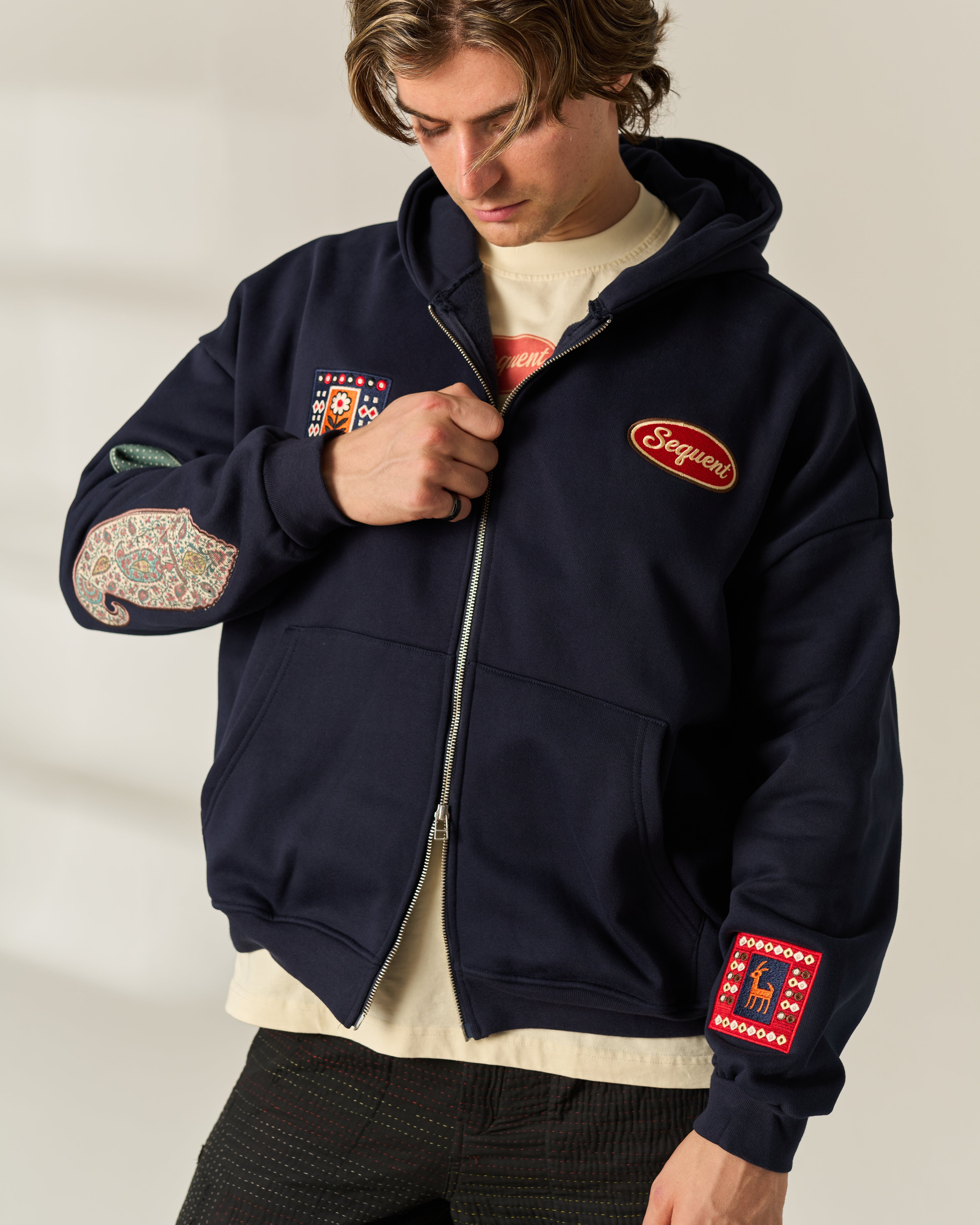 A man wearing a navy blue zip-up hoodie with multiple colorful embroidered patches and a red 'Sequent' logo on the chest, layered over a cream t-shirt and dark pants.