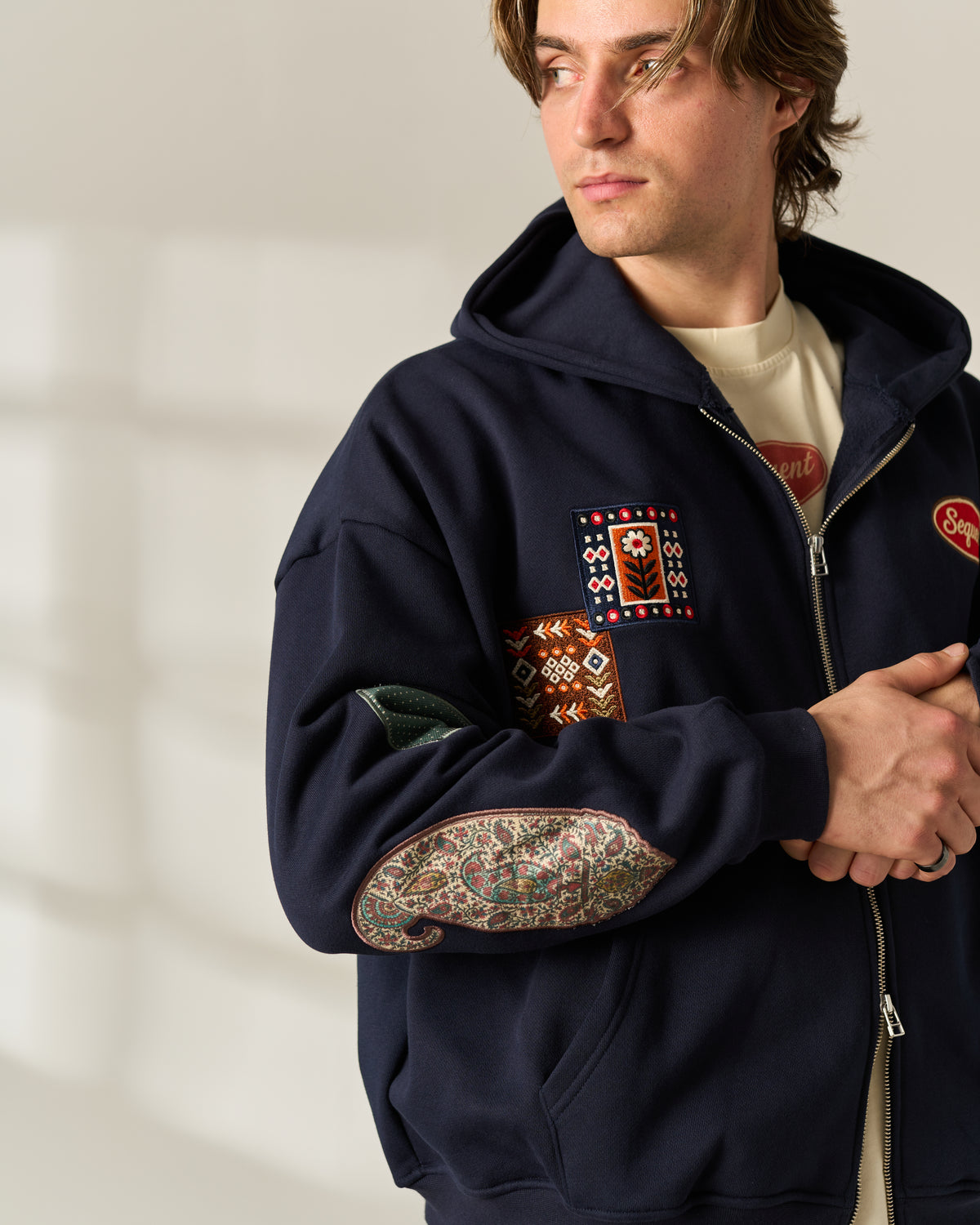 A man wearing a navy blue zip-up hoodie with colorful embroidered patches on the chest and sleeves, styled over a cream t-shirt in a softly lit indoor setting.