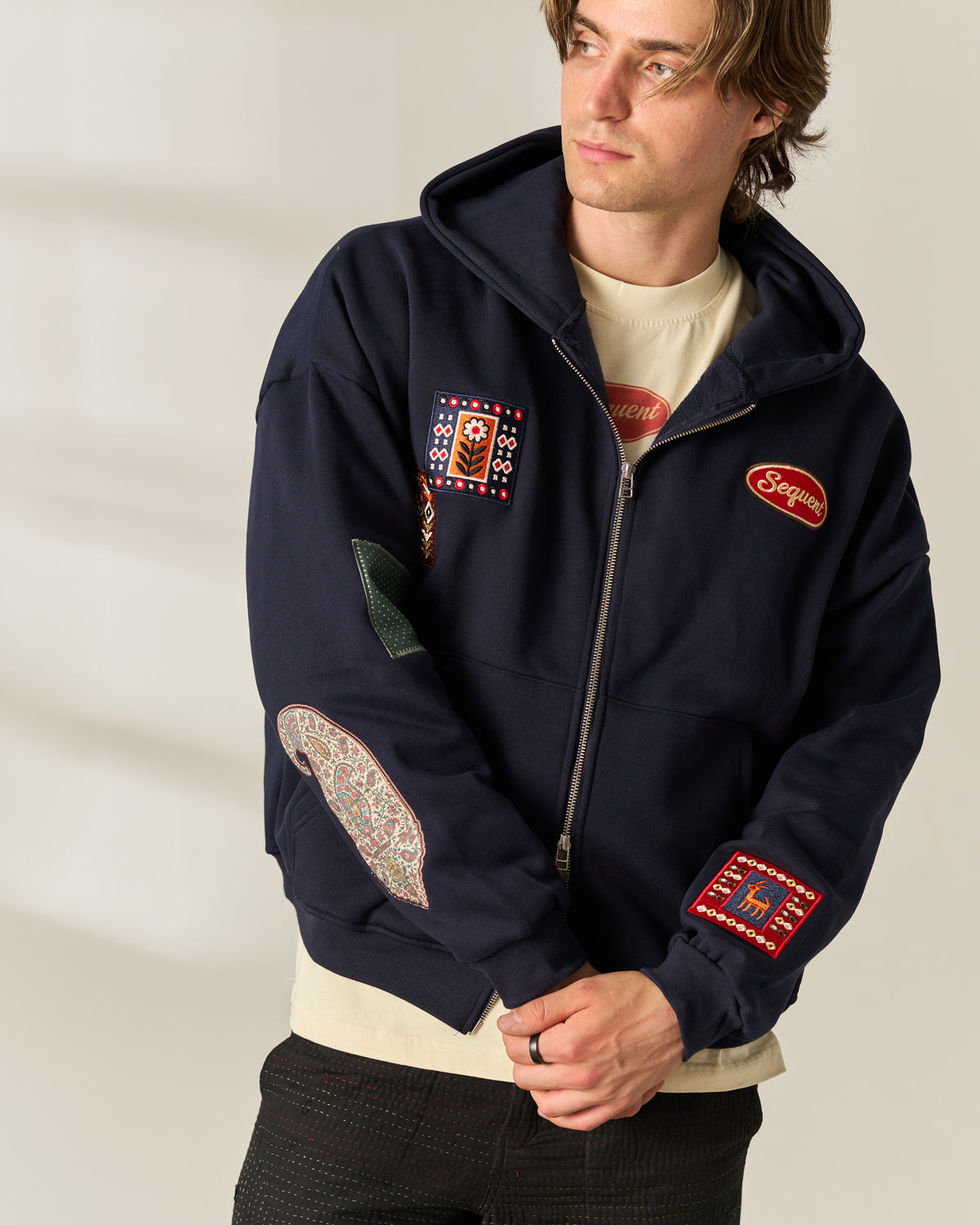 A man wearing a navy blue zip-up hoodie with multiple colorful embroidered patches, layered over a cream t-shirt and dark pants, photographed against a neutral light beige background. The chest has the logo 'Sequent' on it.