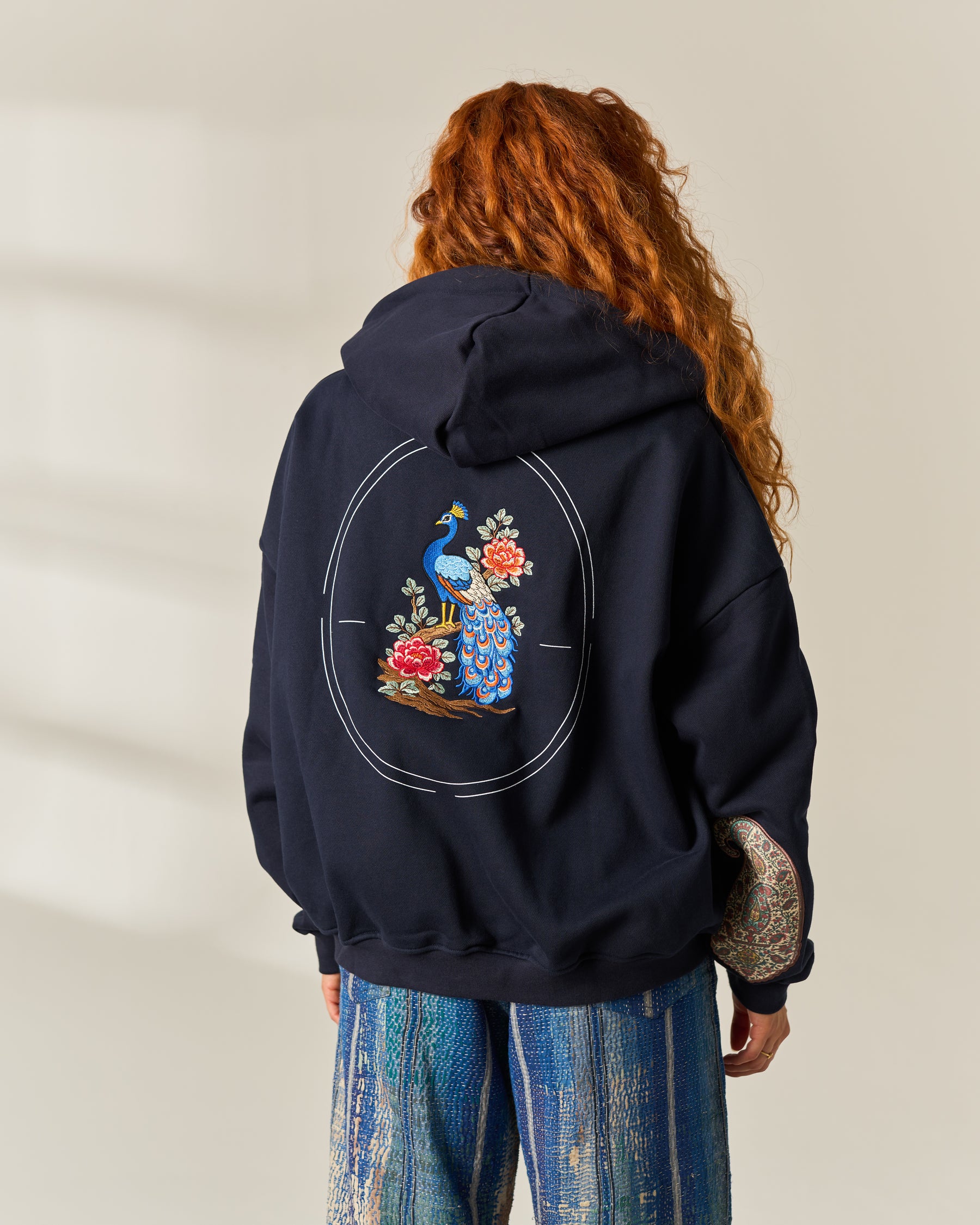 A person viewed from behind wearing a navy hoodie with a large colorful peacock and floral graphic embroidered on the back, paired with blue patterned pants, against a light beige wall.