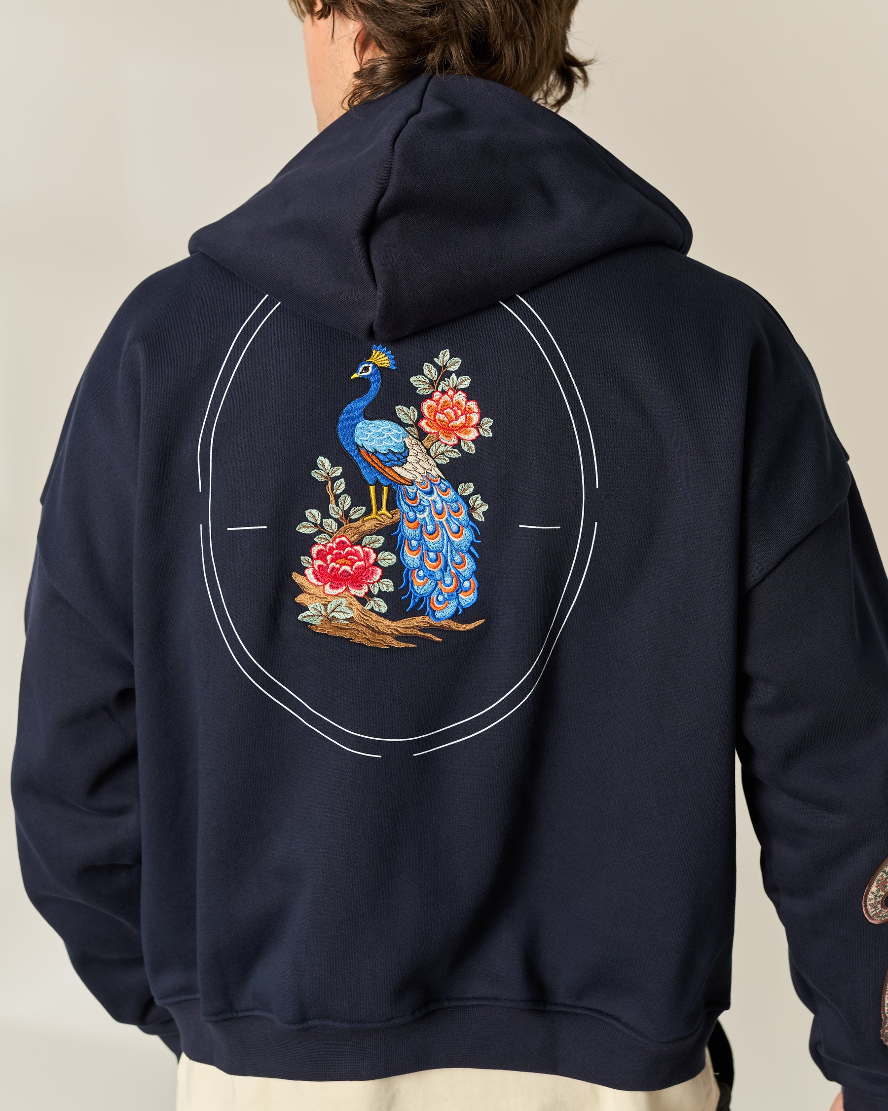A person shown from the back wearing a navy hoodie with a large embroidered peacock and floral design embroidered on the back, set against a plain light beige studio background.