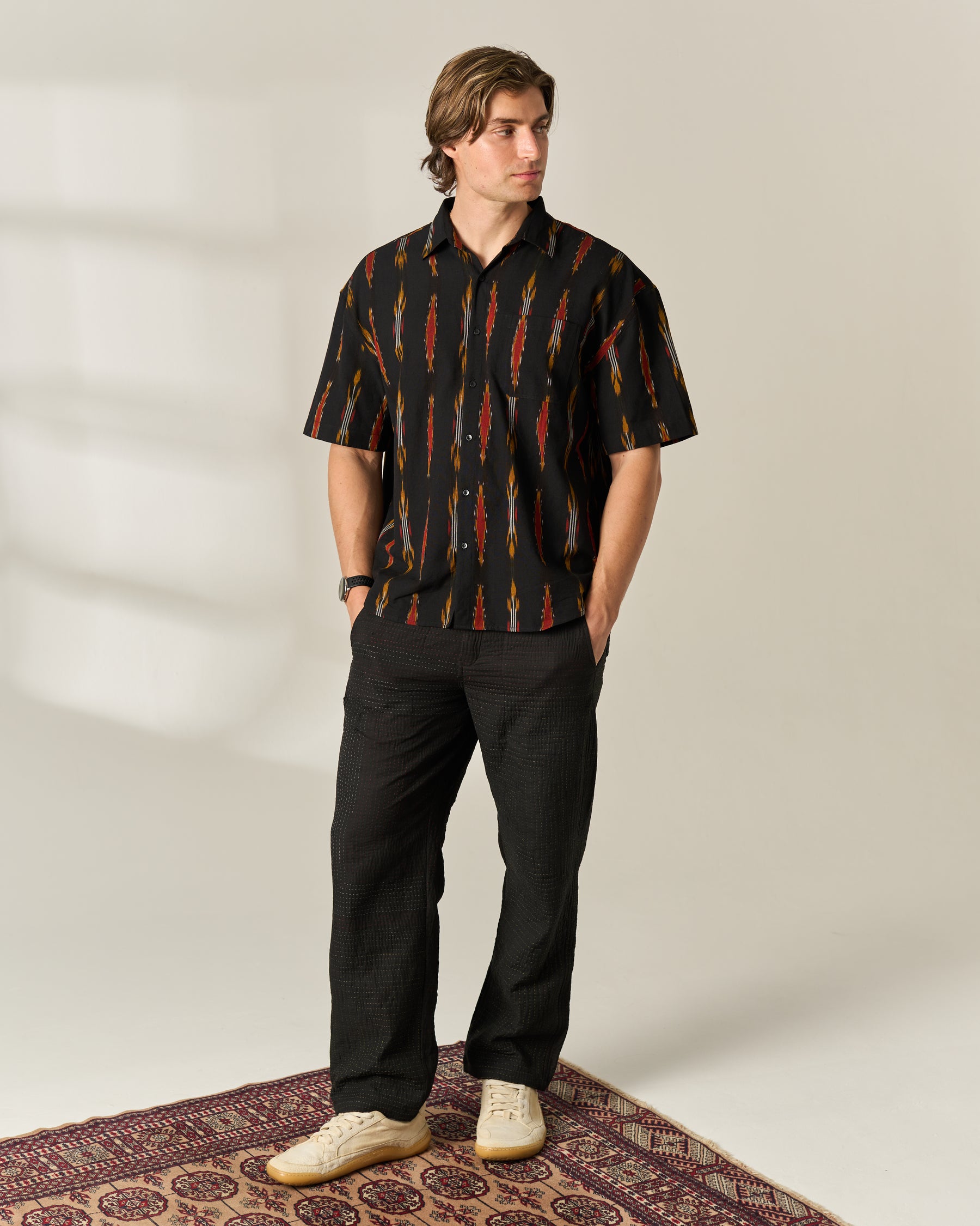 A man stands on a patterned rug wearing a black short-sleeve button-up shirt with a red and orange vertical ikat-style pattern, black trousers, and off-white sneakers in a studio setting.