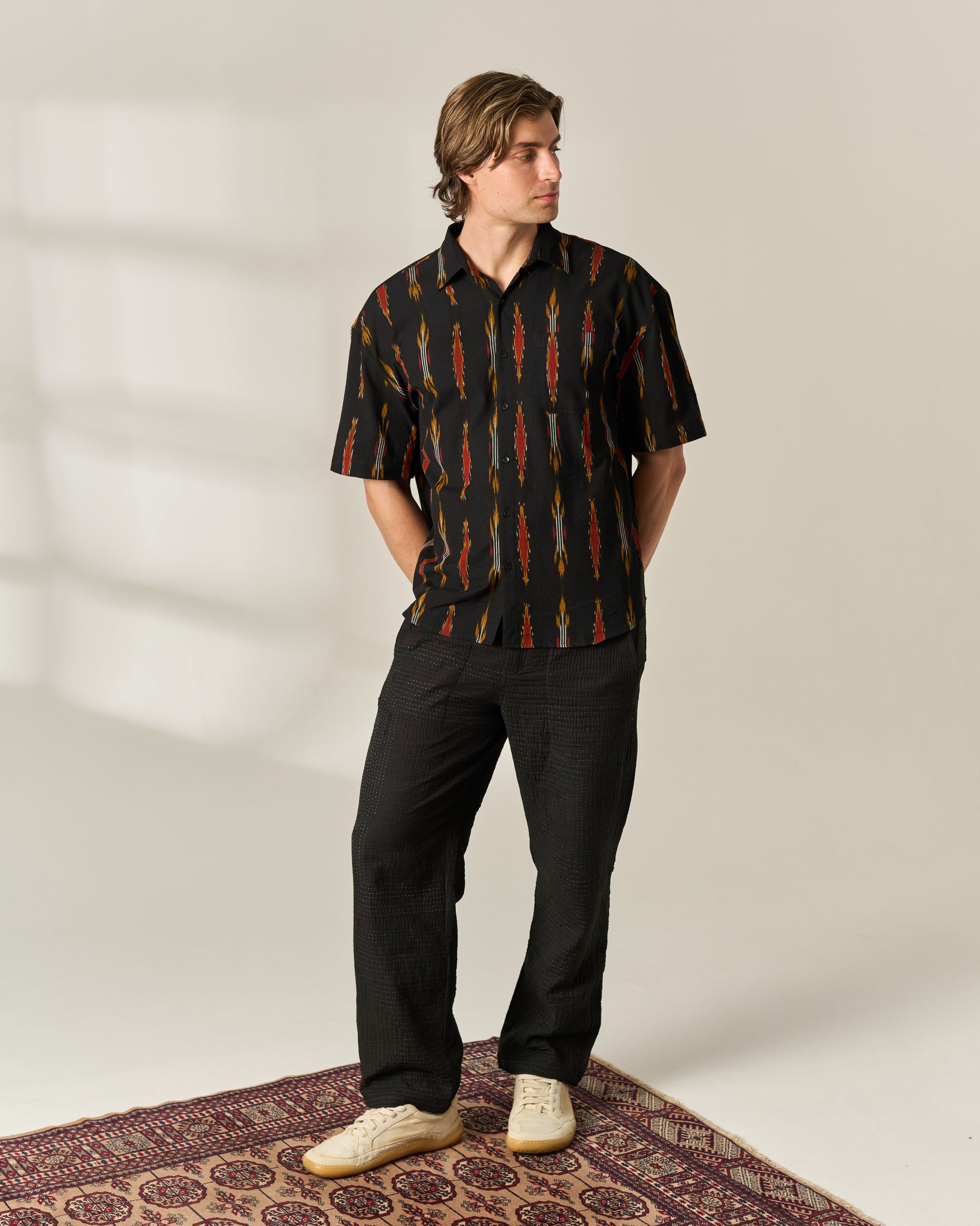 A man stands on a patterned rug wearing a black short-sleeve button-up handwoven Ikat shirt with a vertical multicolor print, black trousers, and off-white sneakers in a brightly lit studio setting.