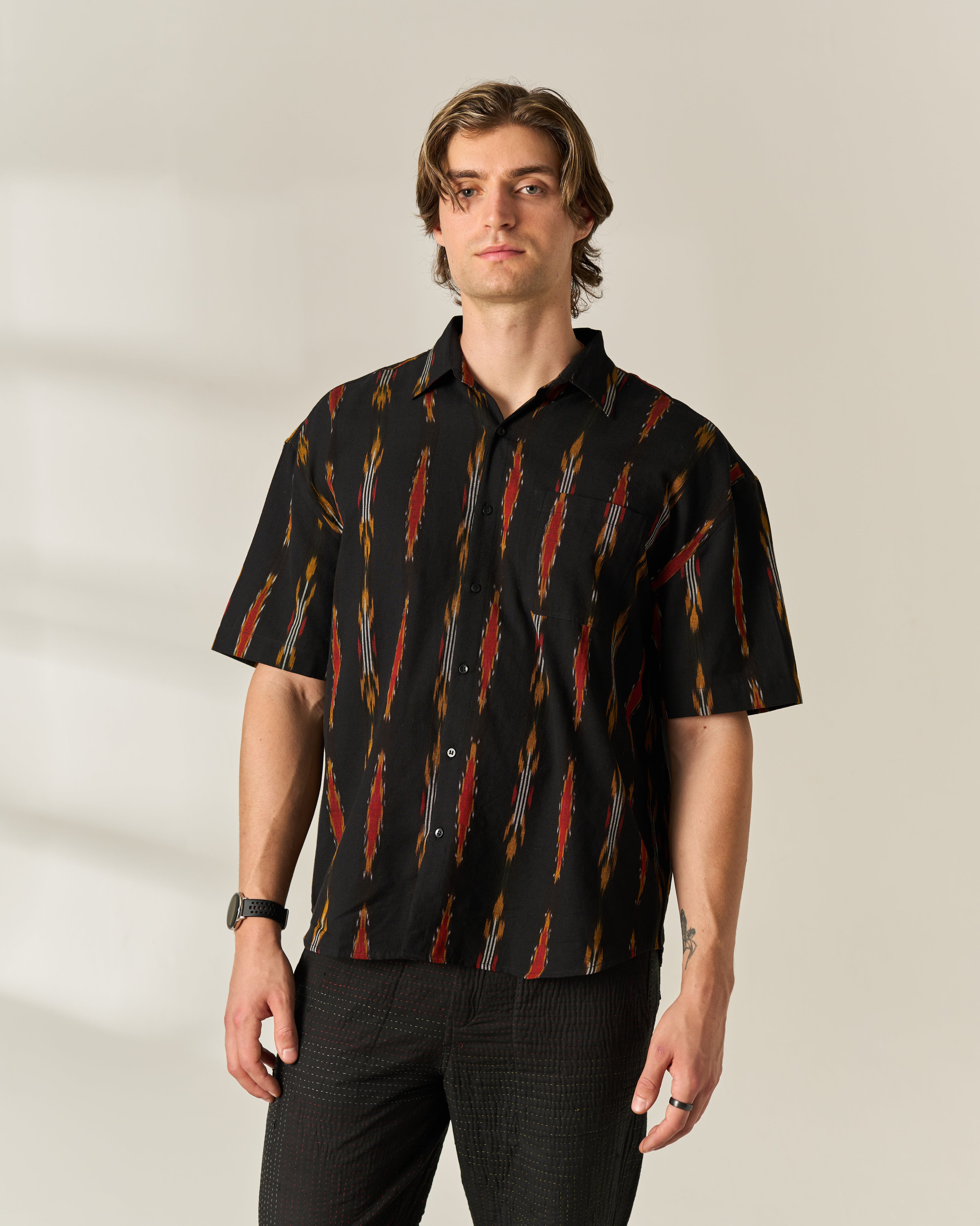 A man wearing a black short-sleeve button-up shirt with a red, orange, and yellow hand woven ikat pattern, paired with dark pants, standing against a neutral beige background.