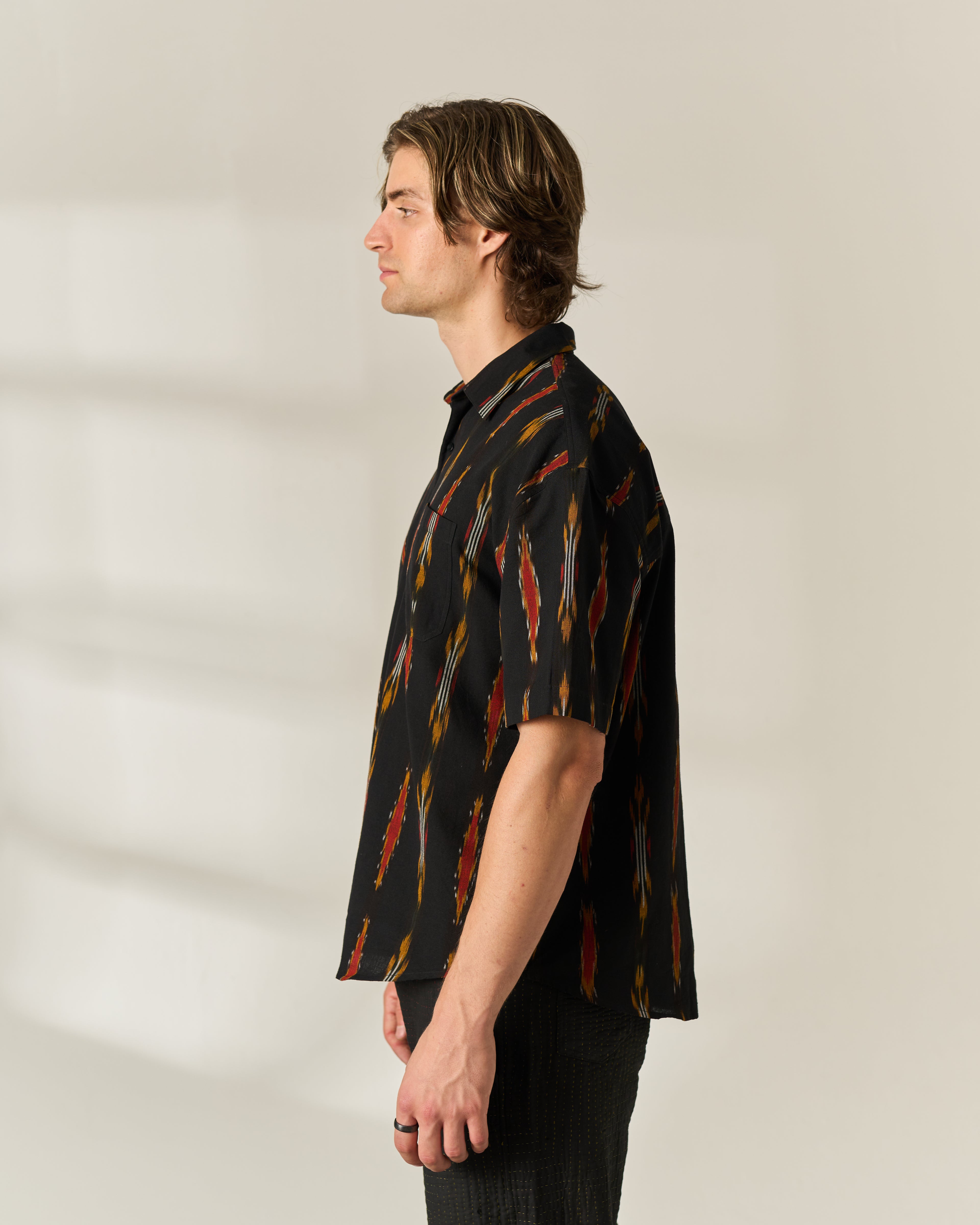A man in profile wearing a short-sleeve button-up shirt with a black base and a vertical multicolor handwoven Ikat pattern in red, orange, and yellow, paired with dark pants against a neutral studio background.