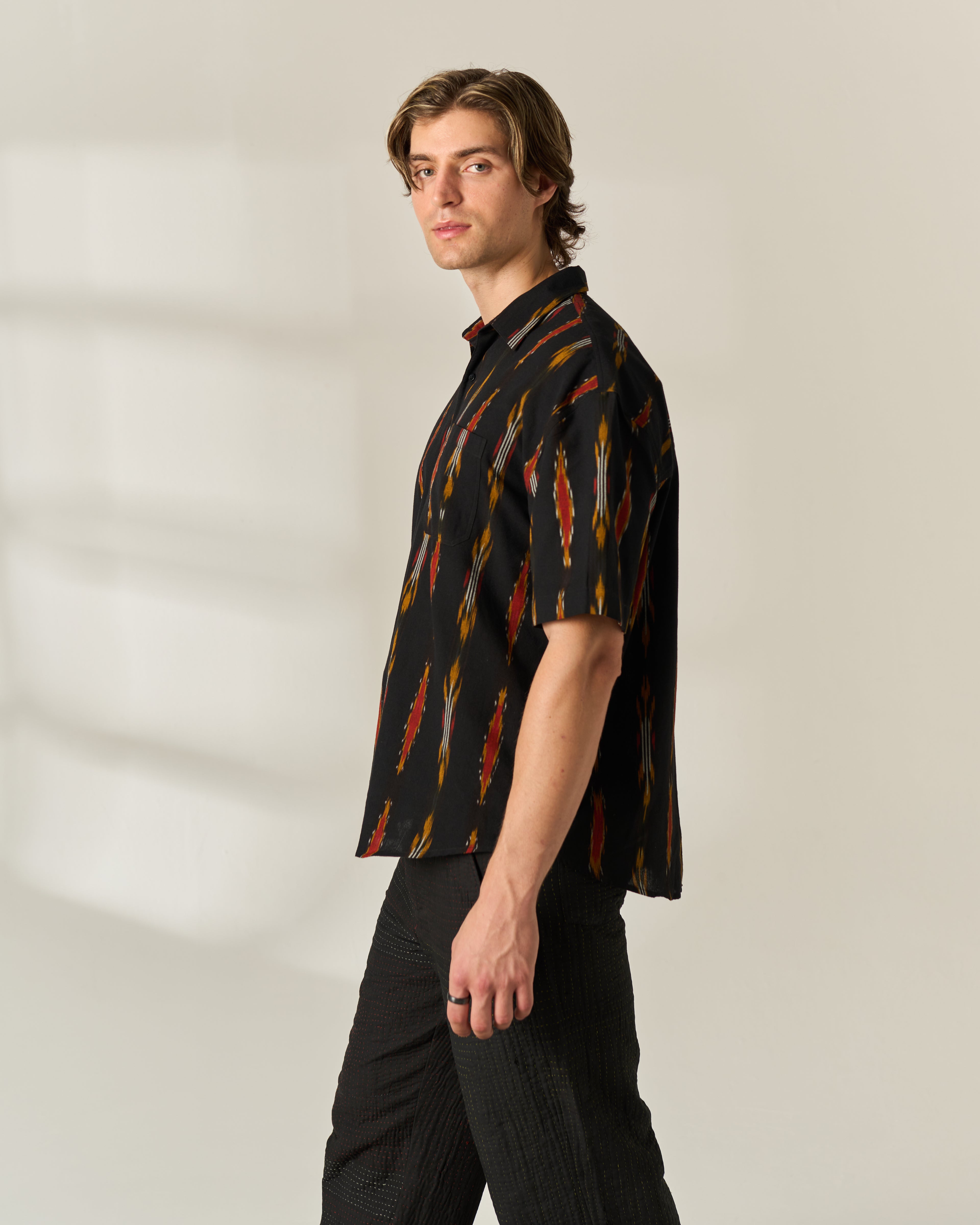 A man wearing a short-sleeve button-up shirt with a black base and vertical red, orange, and yellow feather-like hand woven Ikat patterns, paired with black trousers, standing against a light beige studio background.