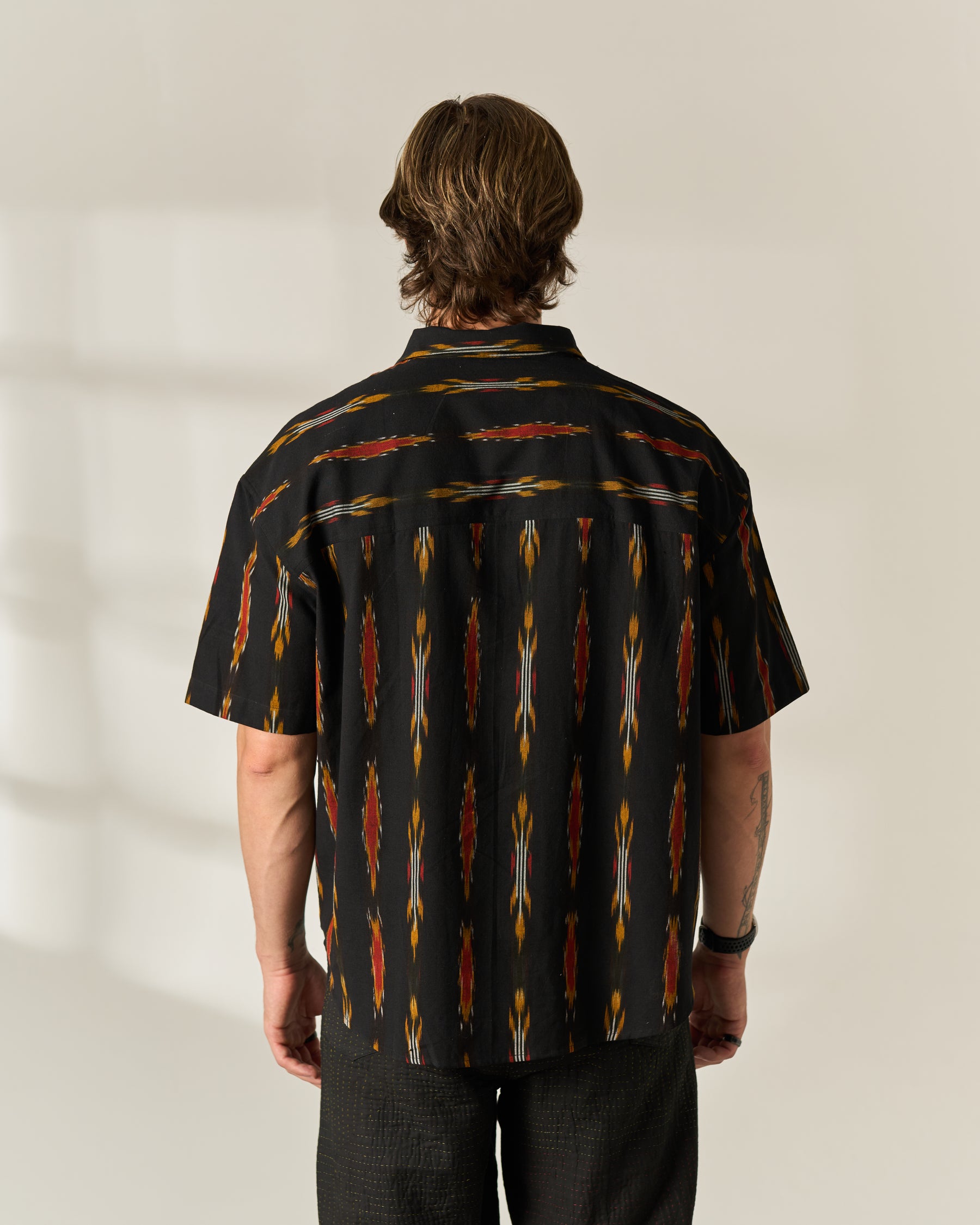 A man viewed from behind wearing a short-sleeve button-up shirt with a black base and a vertical geometric handwoven Ikat pattern in red, orange, and yellow, paired with dark pants against a neutral studio background.