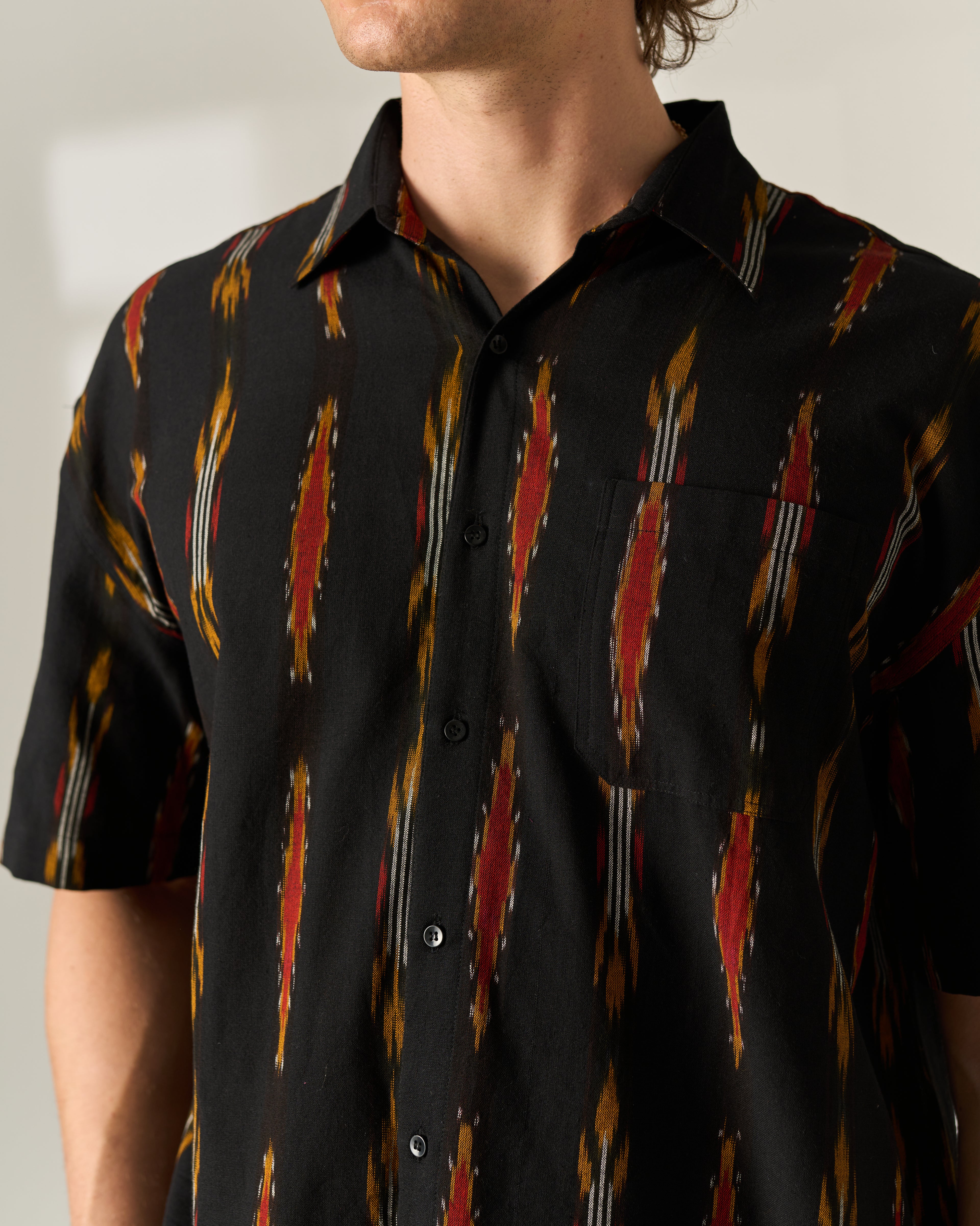 A man wearing a black short-sleeve button-up shirt with a red, orange, and white vertical Handwoven Ikat pattern, shown from the neck down against a neutral light background.