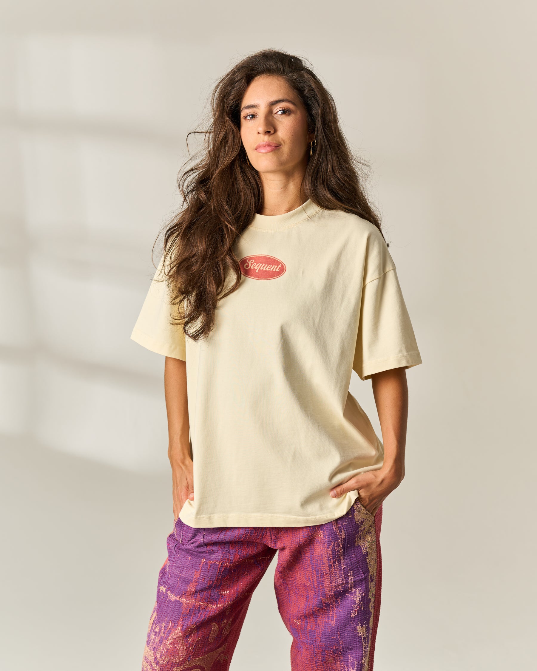 A woman modeling a cream-colored oversized t-shirt with a pink oval 'Sequent' logo on the chest, paired with purple and pink patterned pants, set against a neutral studio background with soft window shadows.
