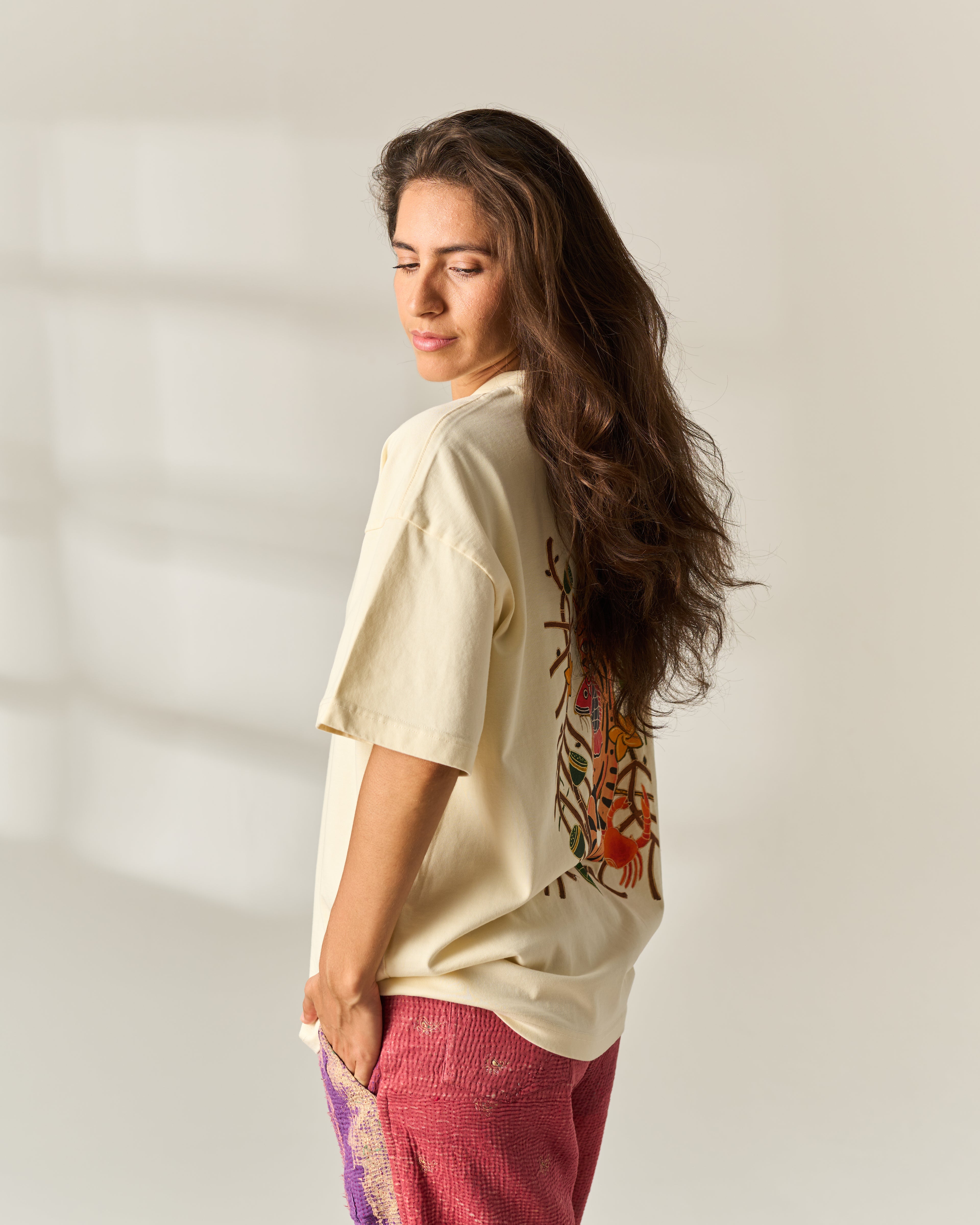 A woman with long dark hair is shown from the side/back wearing an oversized cream-colored t-shirt with a colorful floral graphic on the back, paired with pink patterned pants, set against a minimalist off-white background with soft window shadows.