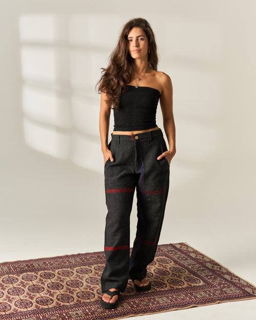 A woman stands on a patterned rug wearing a black strapless tube top and dark wide-leg vintage kantha pants with red and purple embroidered details, photographed against a warm off-white wall with soft window-shadow lighting.