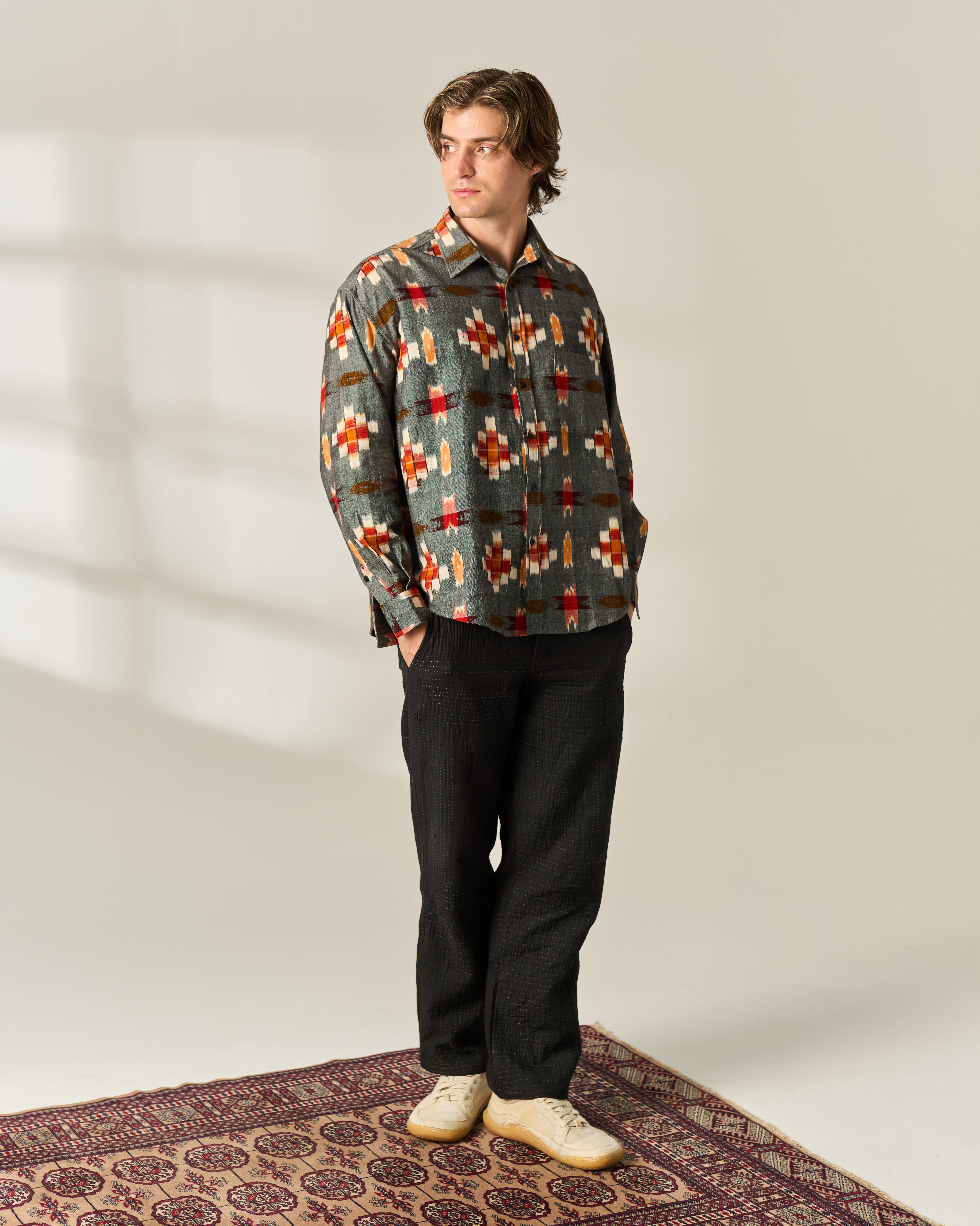 A man stands on a patterned rug wearing a long-sleeve button-up shirt with a muted green base and a colorful geometric handwoven Ikat pattern in orange, red, and beige, paired with dark trousers and cream shoes.