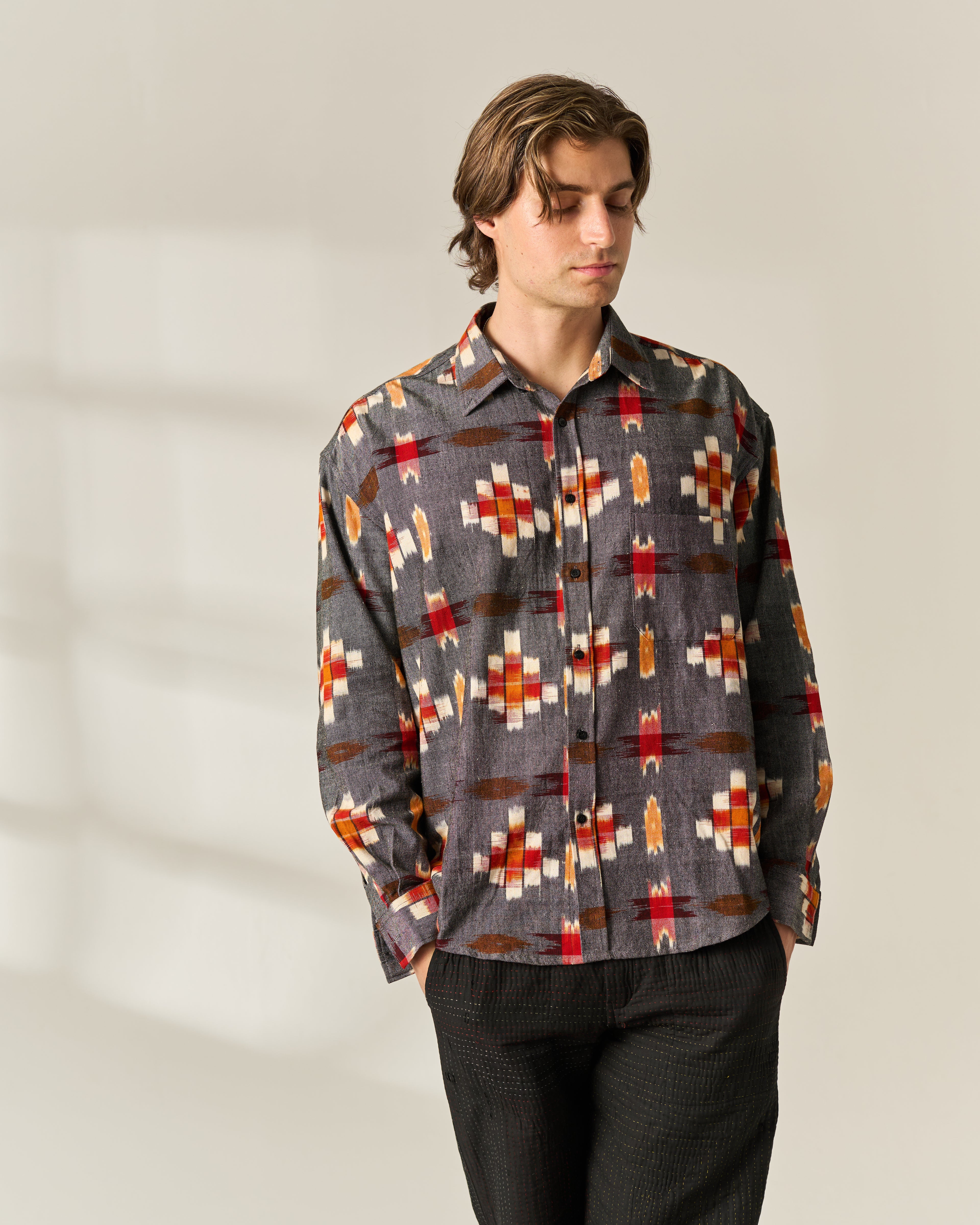 A man wearing a long-sleeve button-up handwoven Ikat shirt with a gray base and a red, orange, and white geometric pattern, paired with dark charcoal pants, standing against a light beige background.