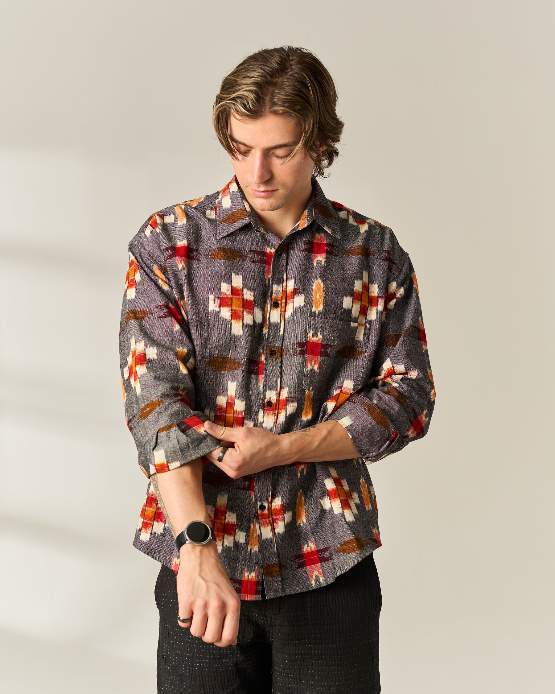 A man wearing a long-sleeve button-up hand woven Ikat shirt with a geometric pattern in gray, red, orange, and cream, paired with black pants, standing against a neutral light gray background.