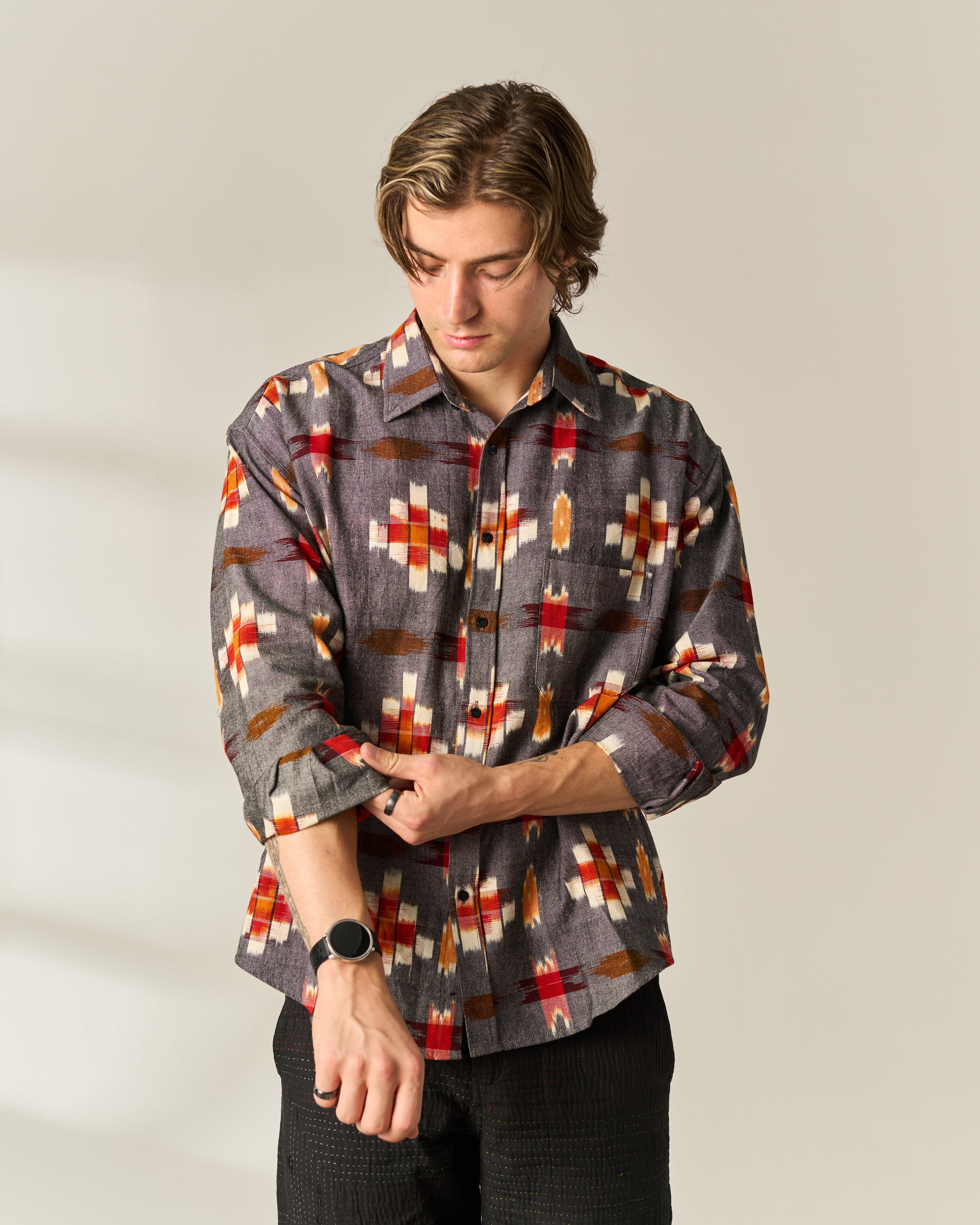 A man wearing a long-sleeve button-up hand woven Ikat shirt with a geometric pattern in gray, red, orange, and cream, paired with black pants, standing against a neutral light gray background.