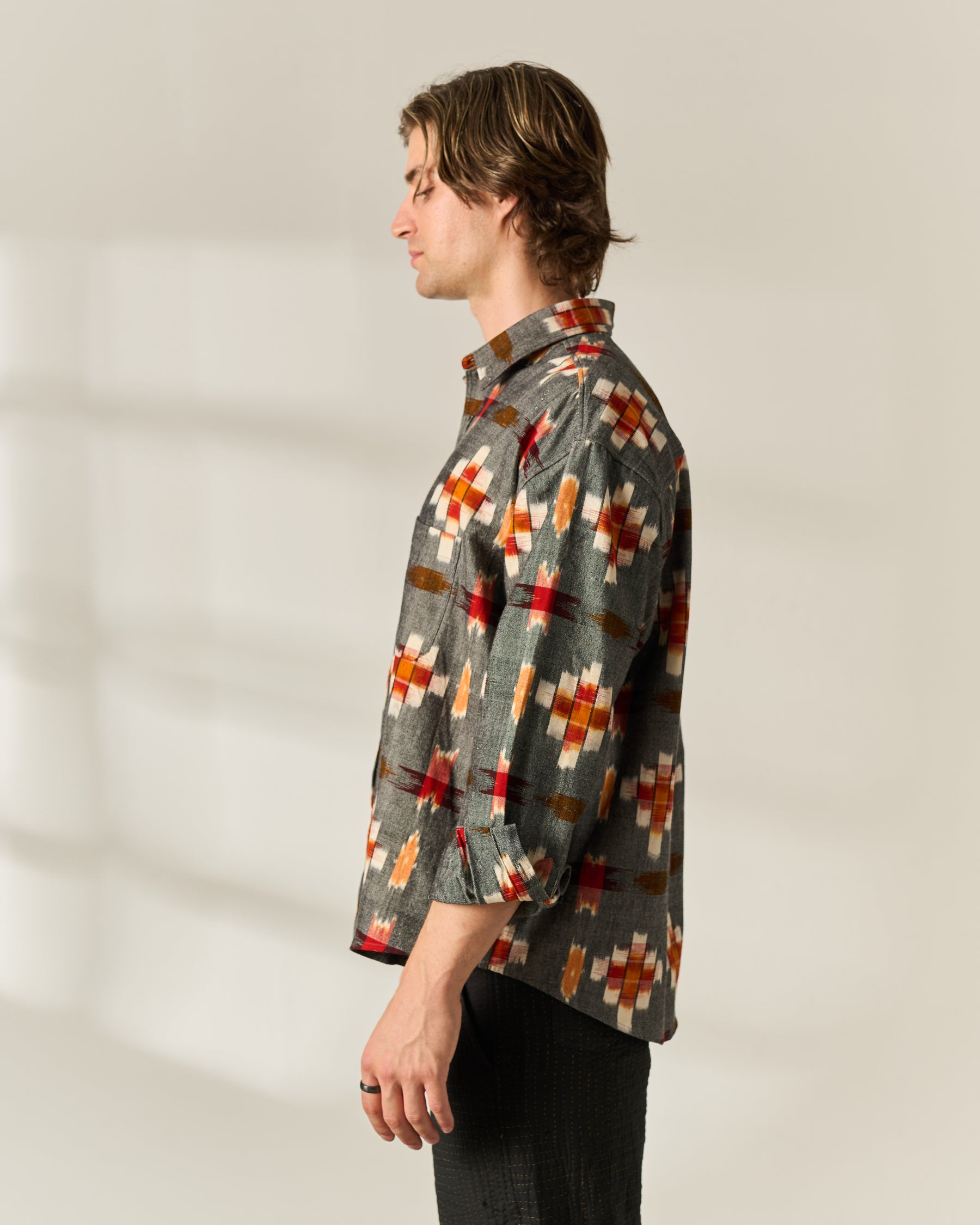 A man shown in profile wearing a long-sleeve button-up handwoven Ikat shirt with a gray base and a geometric pattern in red, orange, brown, and white, paired with dark trousers against a warm off-white background.