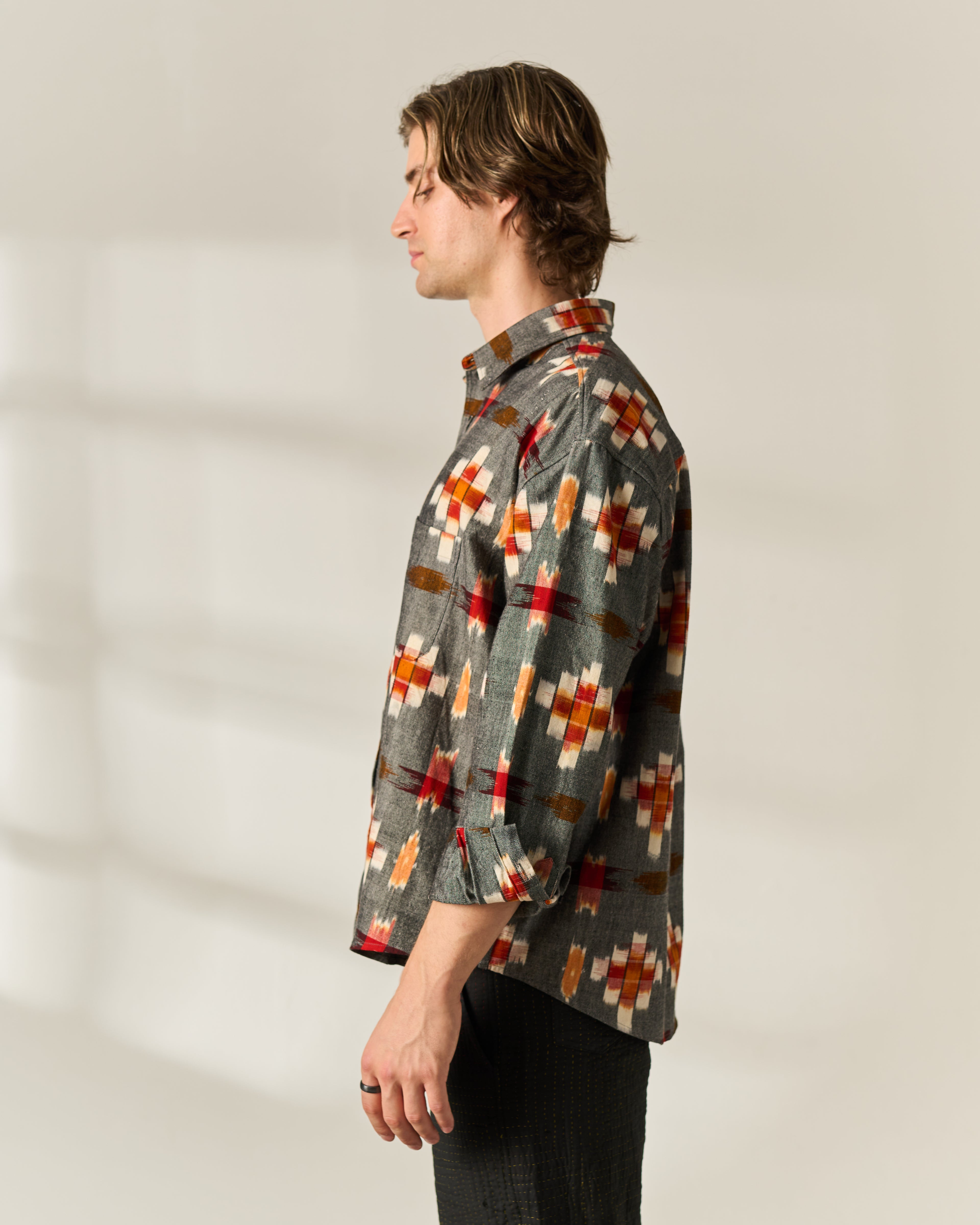 A man shown in profile wearing a long-sleeve button-up handwoven Ikat shirt with a gray base and a geometric pattern in red, orange, brown, and white, paired with dark trousers against a warm off-white background.