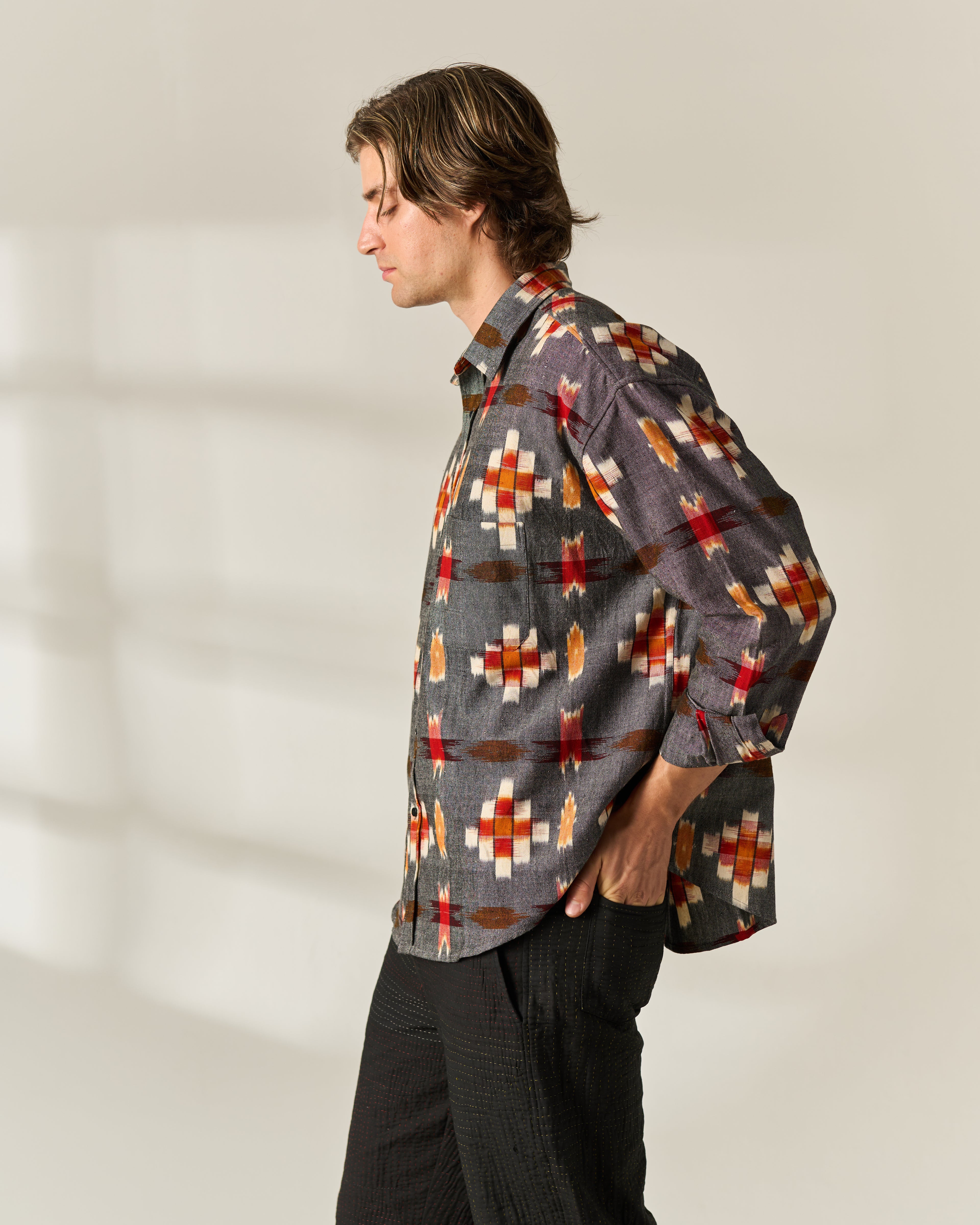 A man wearing a long-sleeve button-up hand woven Ikat shirt with a gray base and a red, orange, and white geometric pattern, paired with dark pants, standing in profile against a warm off-white background with soft window shadows.