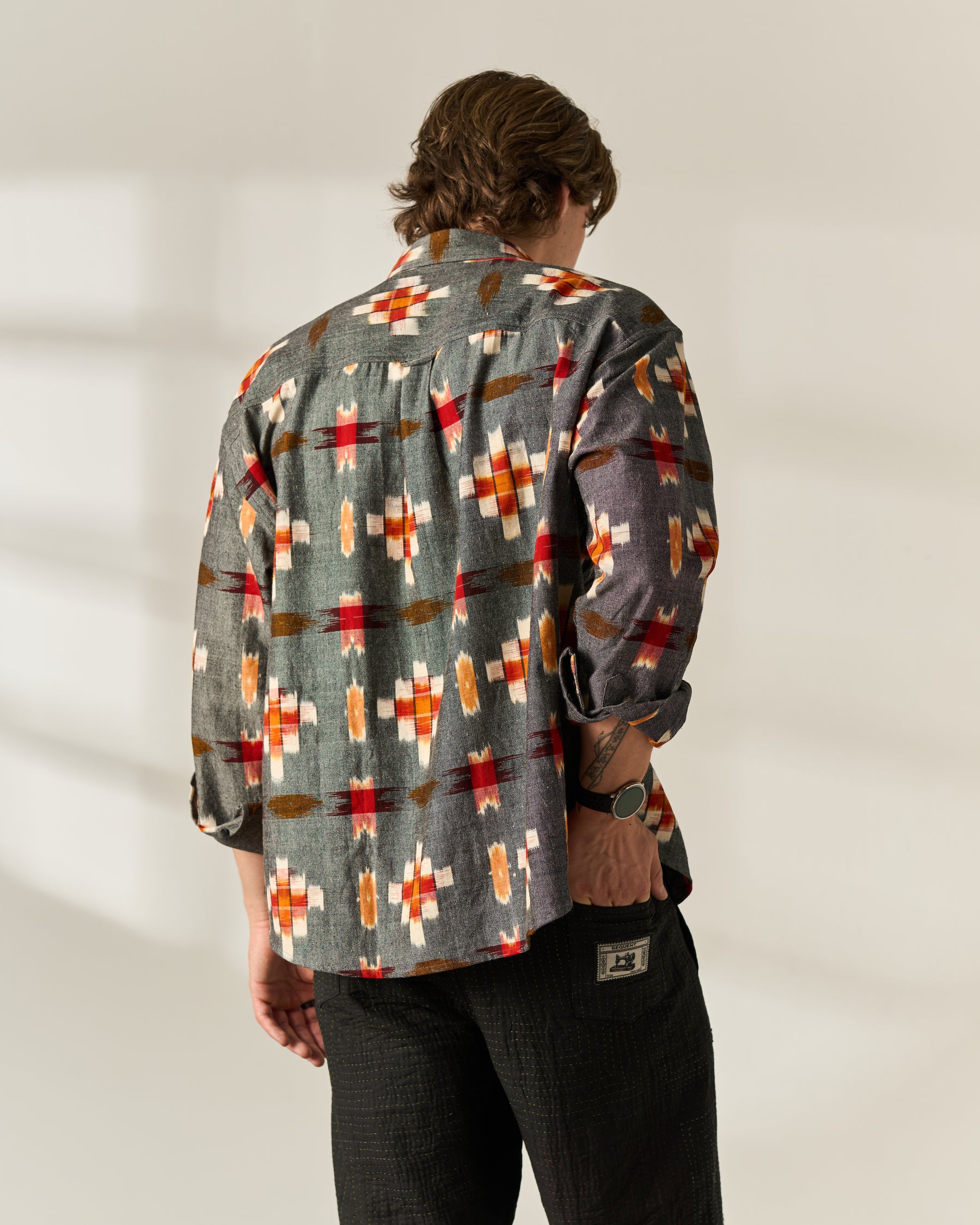 A man viewed from behind wearing a gray long-sleeve button-up handwoven Ikat shirt with a red, orange, and white geometric pattern, paired with black pants, against a warm off-white background.
