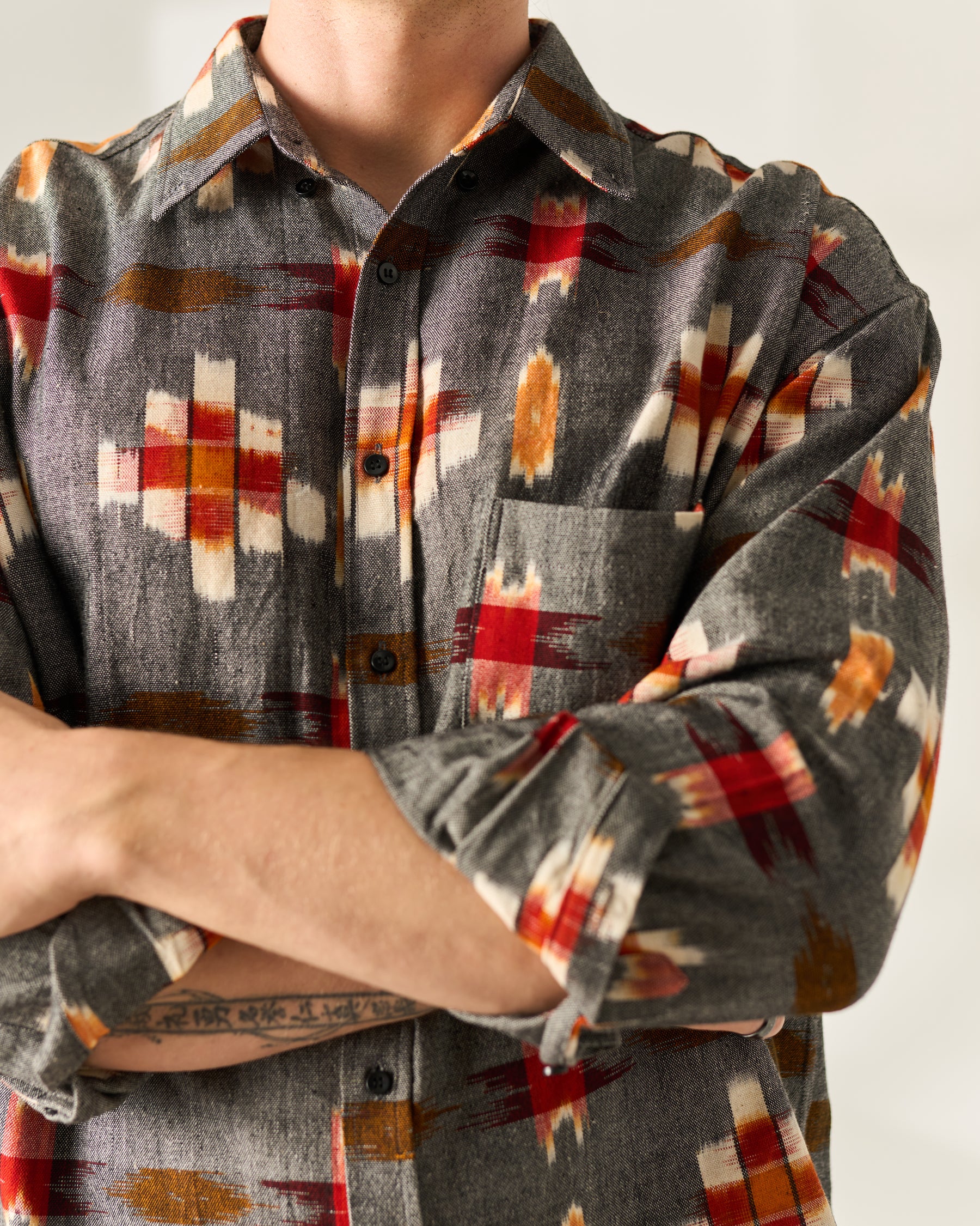A close-up of a person wearing a long-sleeve button-up handwoven Ikat shirt with a gray base and a geometric pattern in red, orange, and cream.