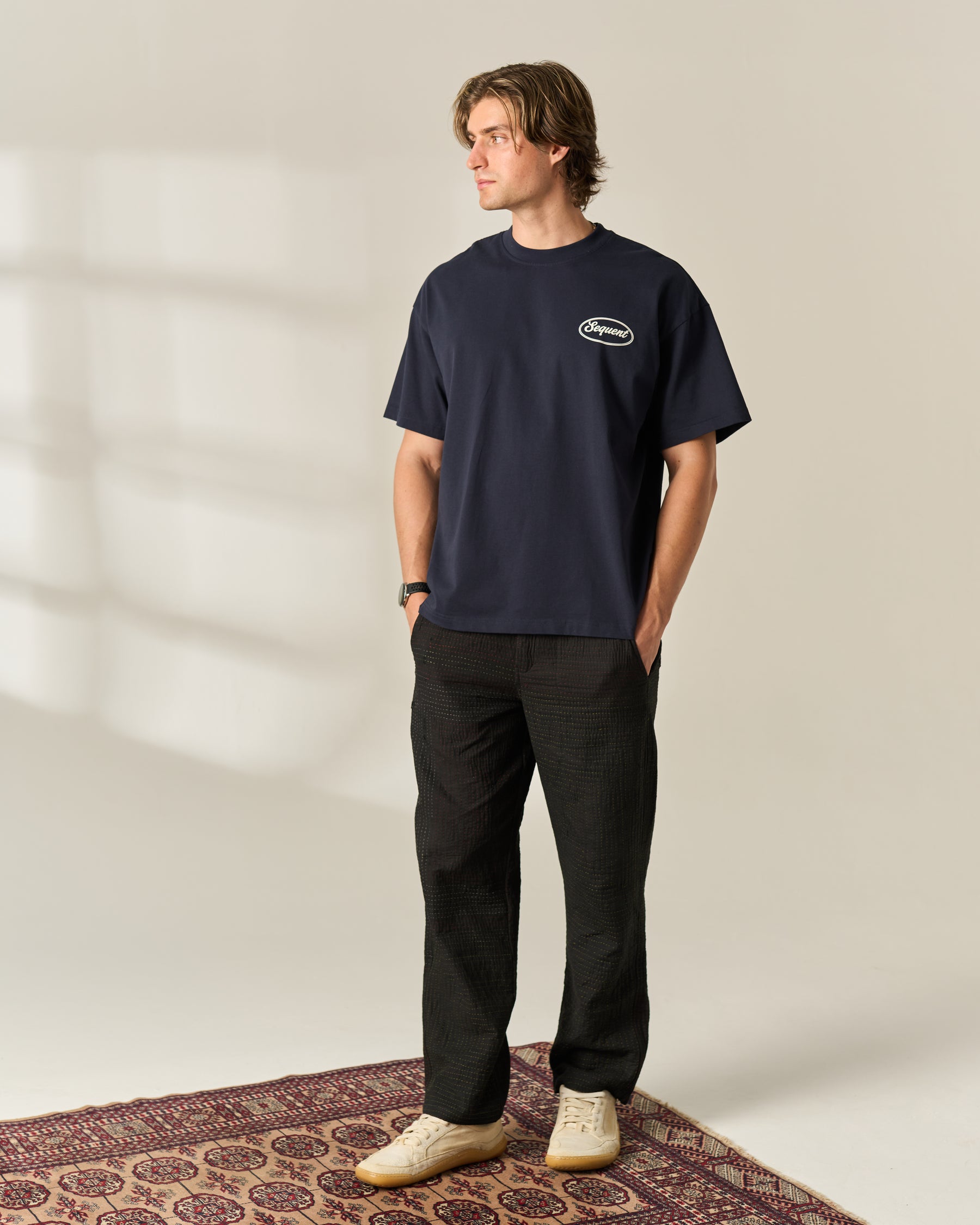 A man wearing a navy blue crewneck t-shirt with a small white oval 'Sequent' logo on the left chest, paired with black trousers and beige sneakers, standing on a patterned rug against a warm beige background.