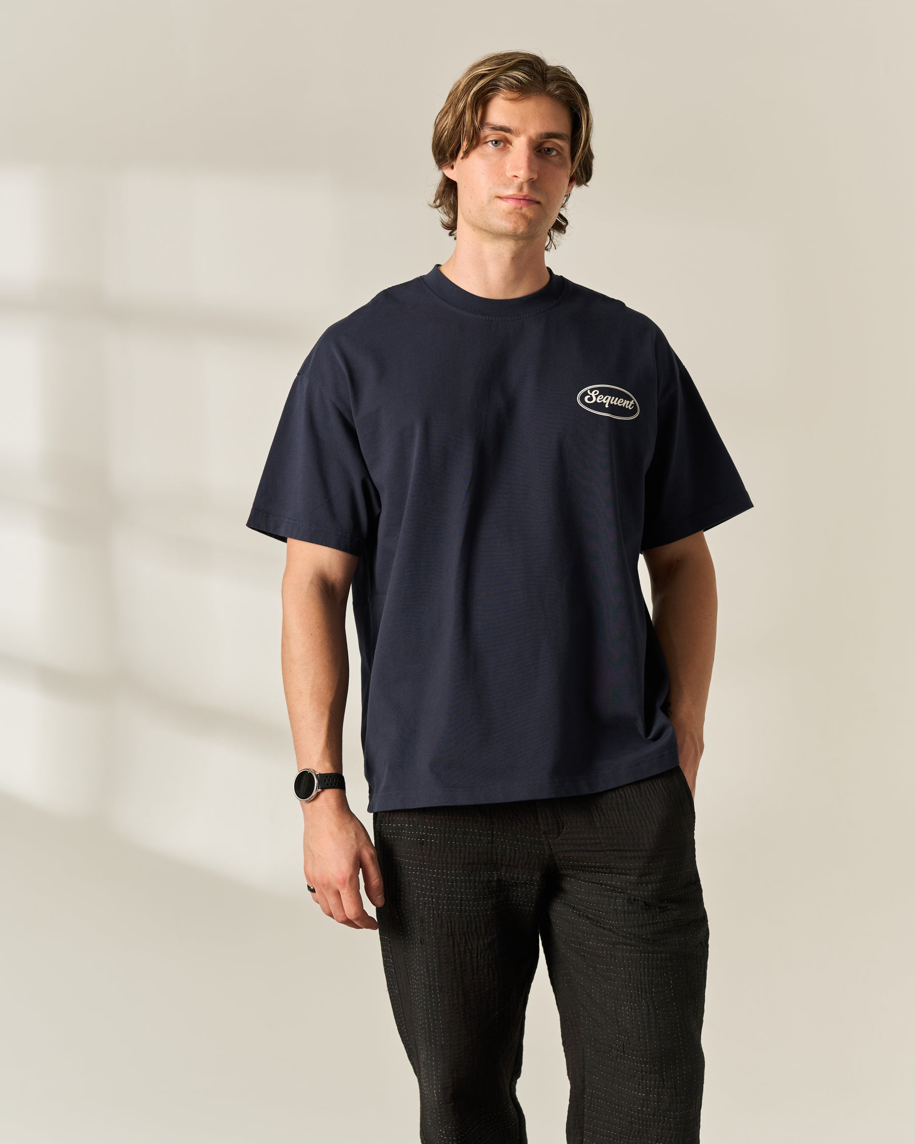 A man wearing a navy blue crewneck t-shirt with a small white oval 'Sequent' logo on the left chest, paired with dark charcoal pants, standing against a warm beige wall with soft window shadows.