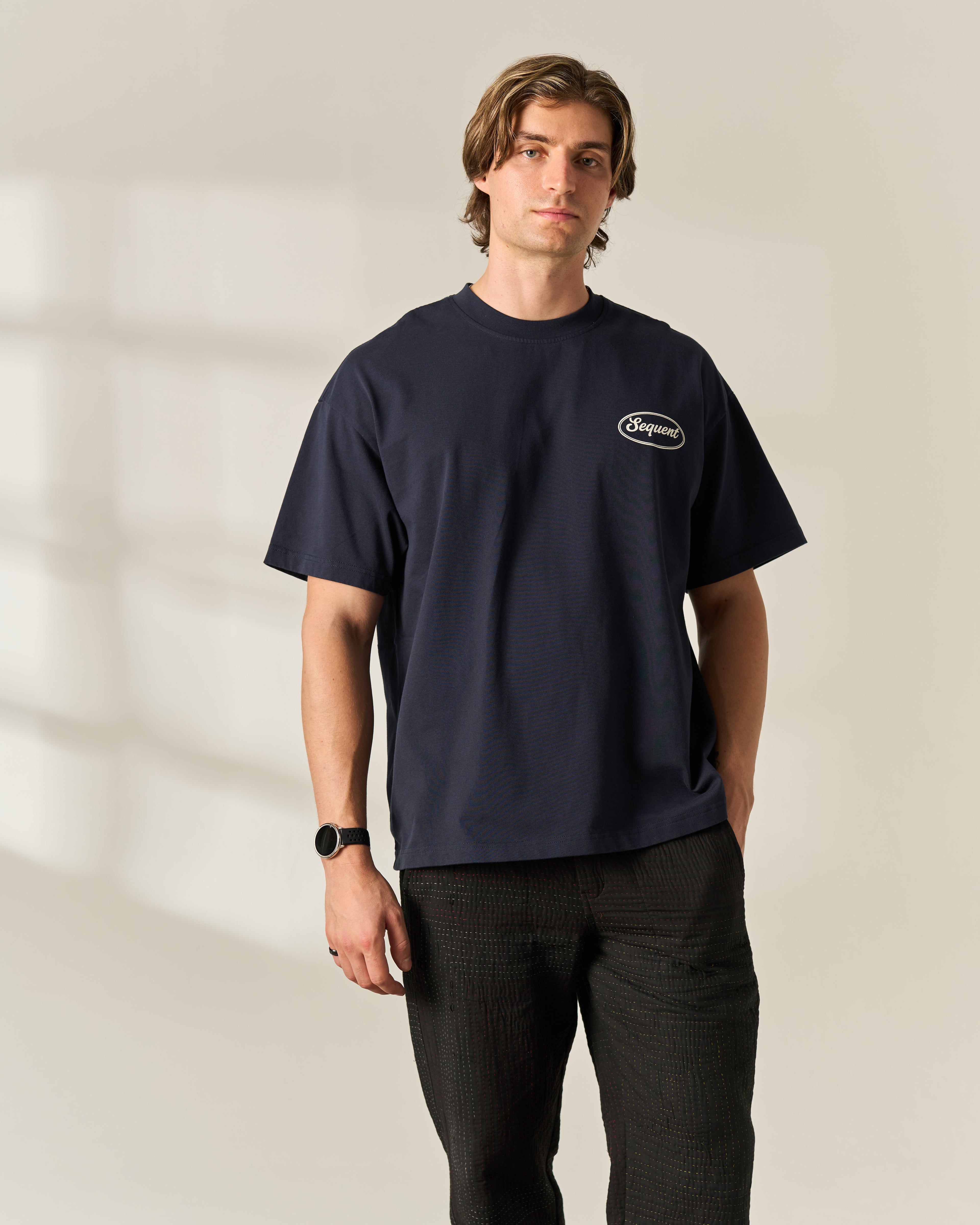 A man wearing a navy blue crewneck t-shirt with a small white oval 'Sequent' logo on the left chest, paired with dark charcoal pants, standing against a warm beige wall with soft window shadows.