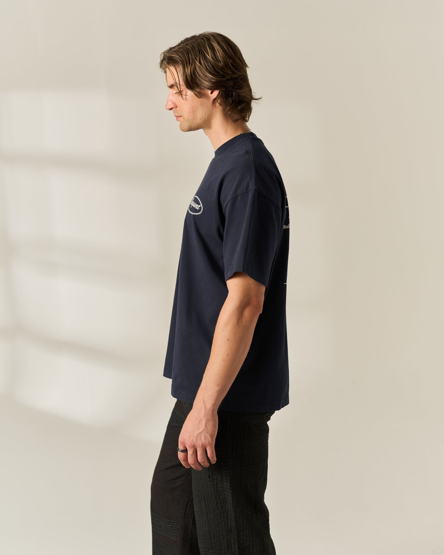 A man in profile wearing a navy blue t-shirt with a small white graphic on the left chest, paired with black pants, standing against a warm off-white background.