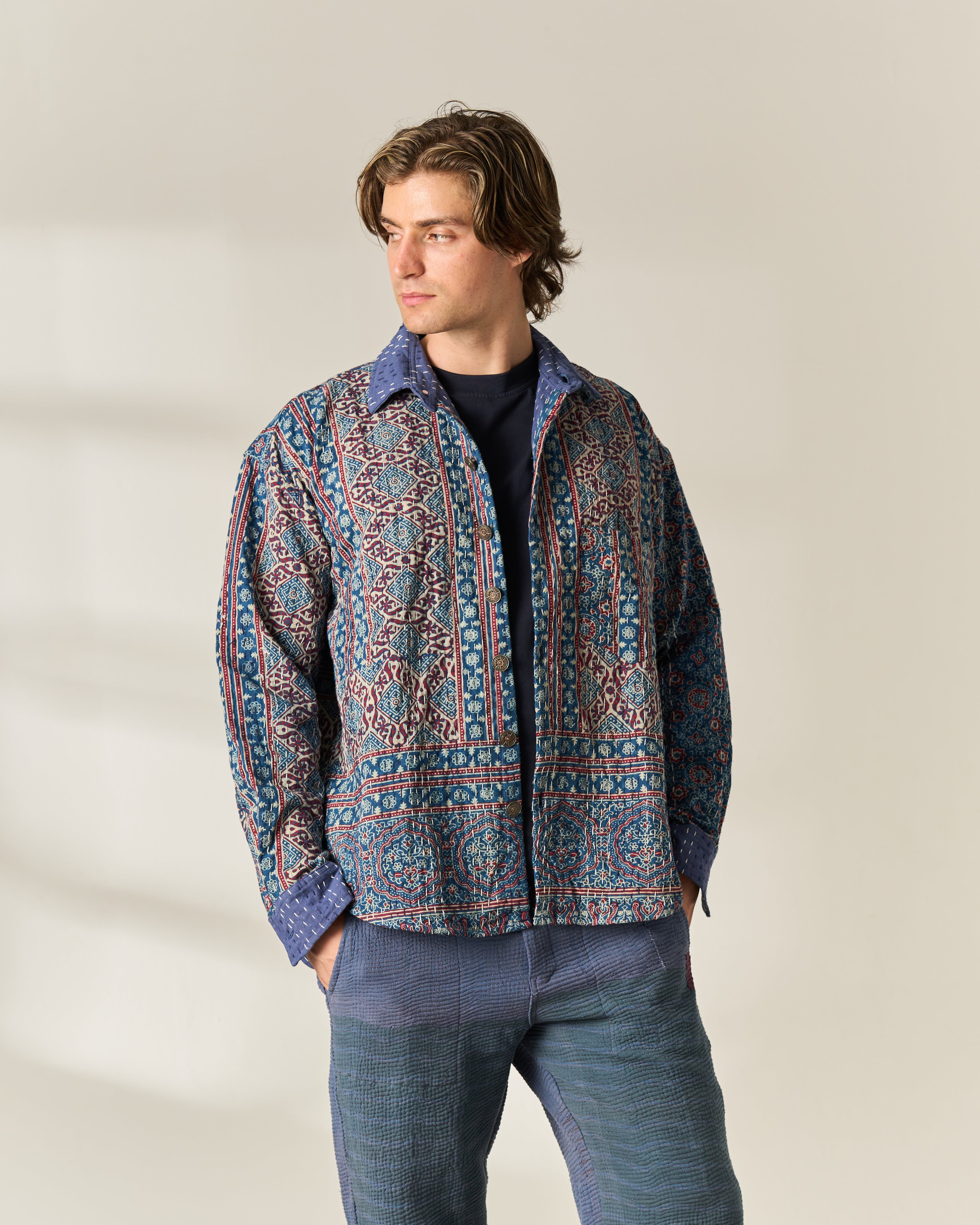 A man wearing a blue and red hand painted Ajrakh Kantha button-up shirt jacket over a black t-shirt and blue trousers, standing against a light beige background.