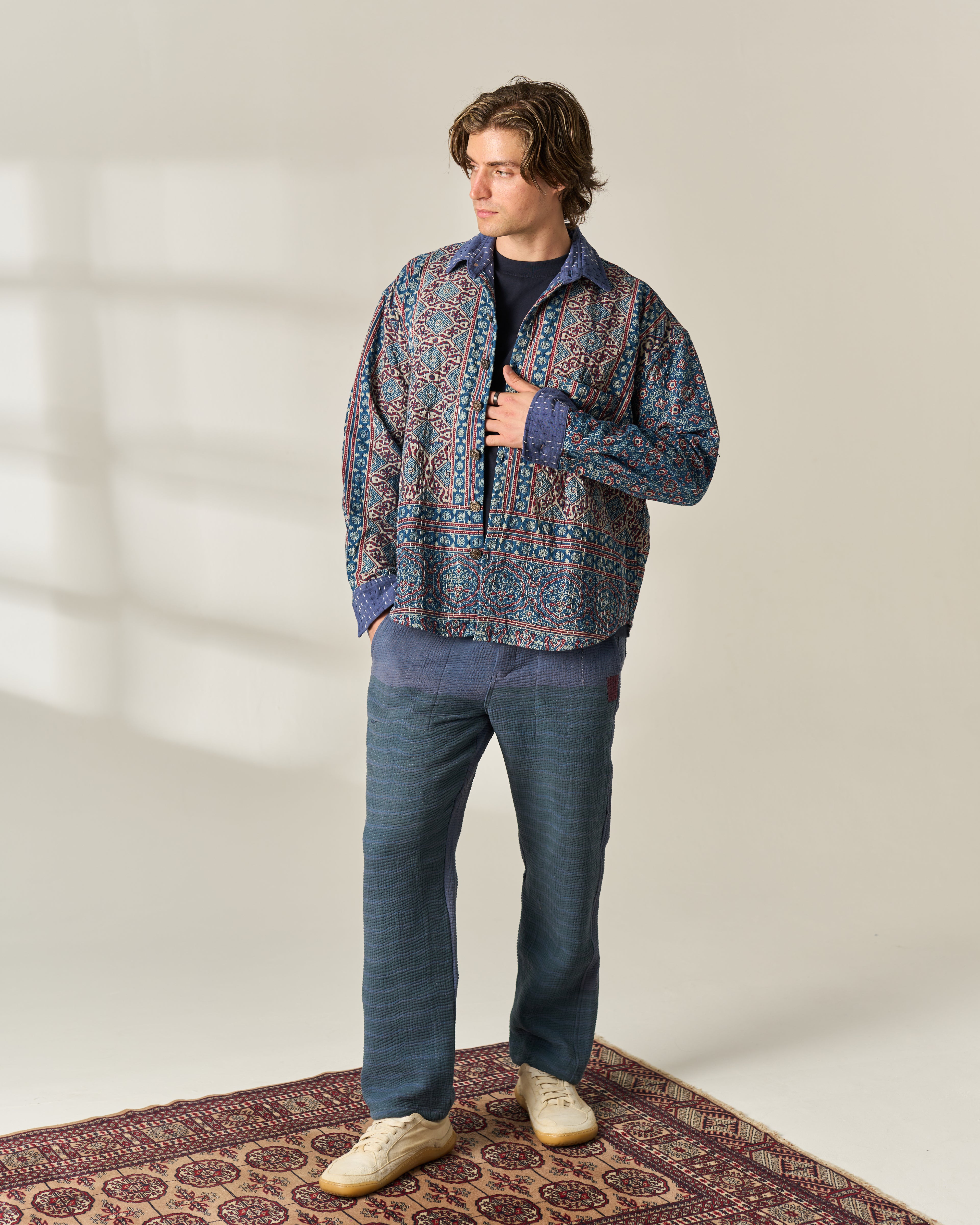 A man stands on a patterned rug wearing a multicolor patterned button-up Ajrakh Kantha shirt over a dark t-shirt, blue jeans, and cream sneakers in a brightly lit studio setting.