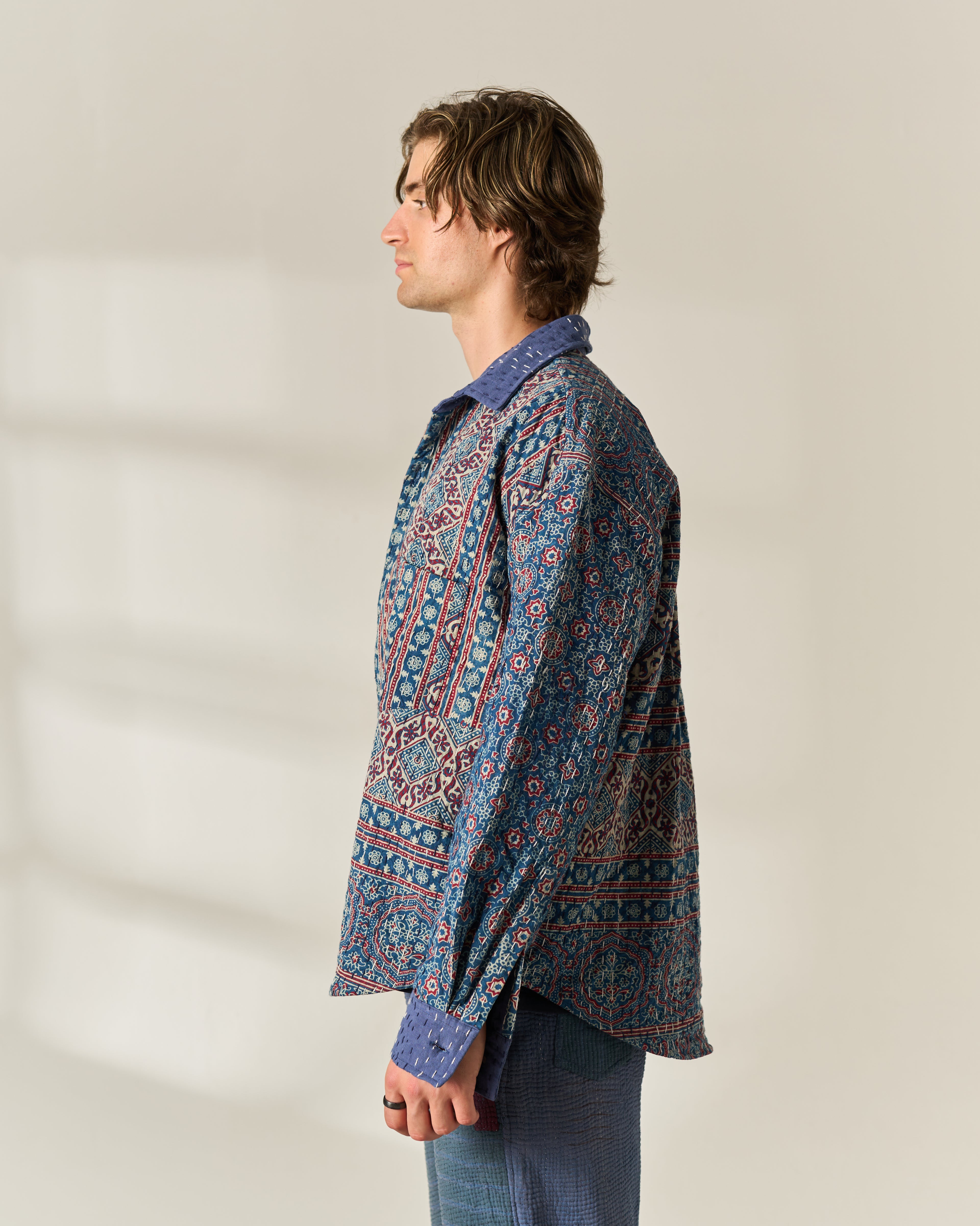 A man in profile wearing a long-sleeve button-up shirt with a detailed blue, red, and white geometric Ajrakh Kantha pattern, paired with blue trousers, set against a warm off-white background with soft shadows.
