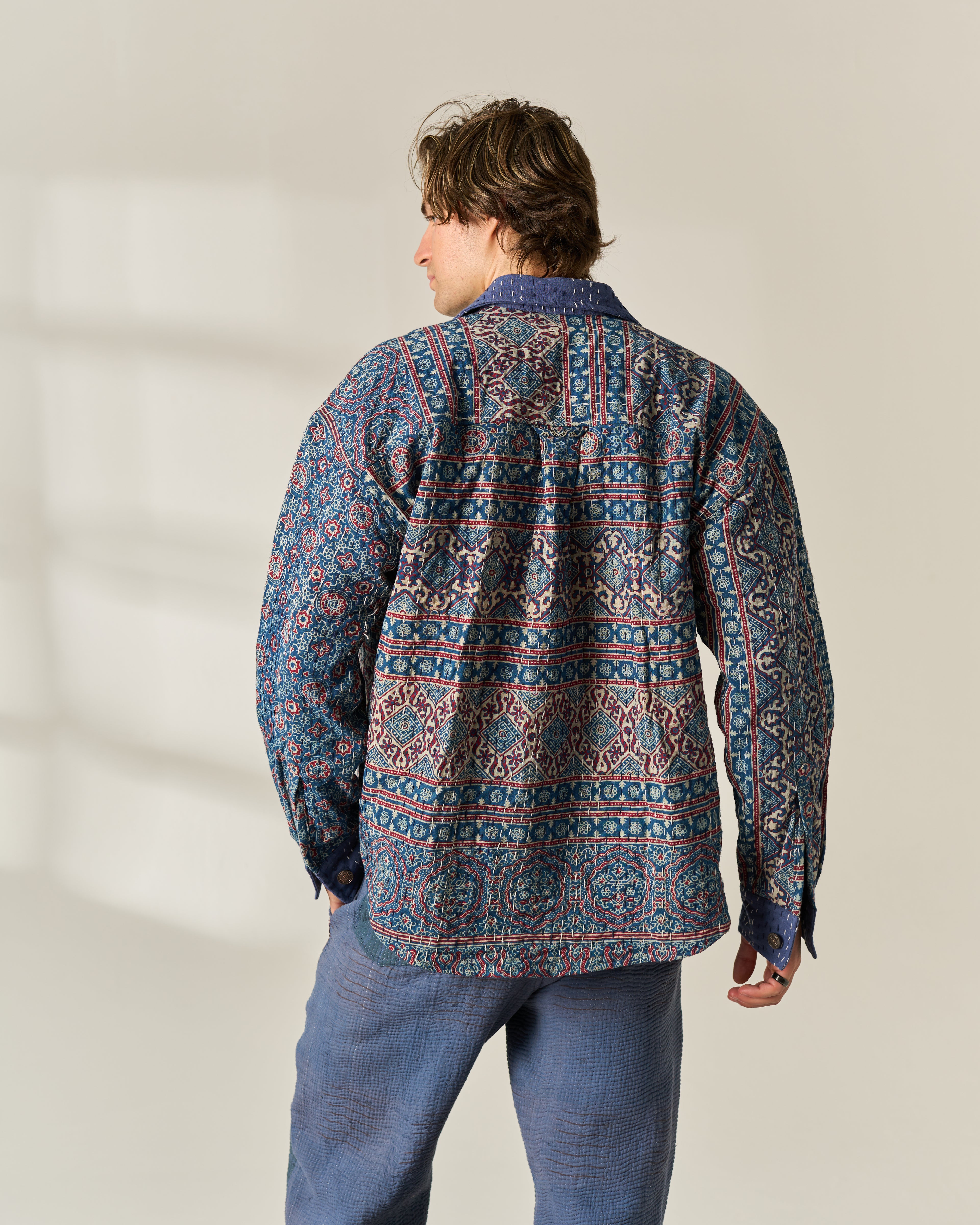 A man viewed from behind wearing a long-sleeve button-up shirt with an intricate multicolor geometric hand block painted Ajrakh Kantha in blue, red, and beige, paired with blue jeans against a neutral beige background.