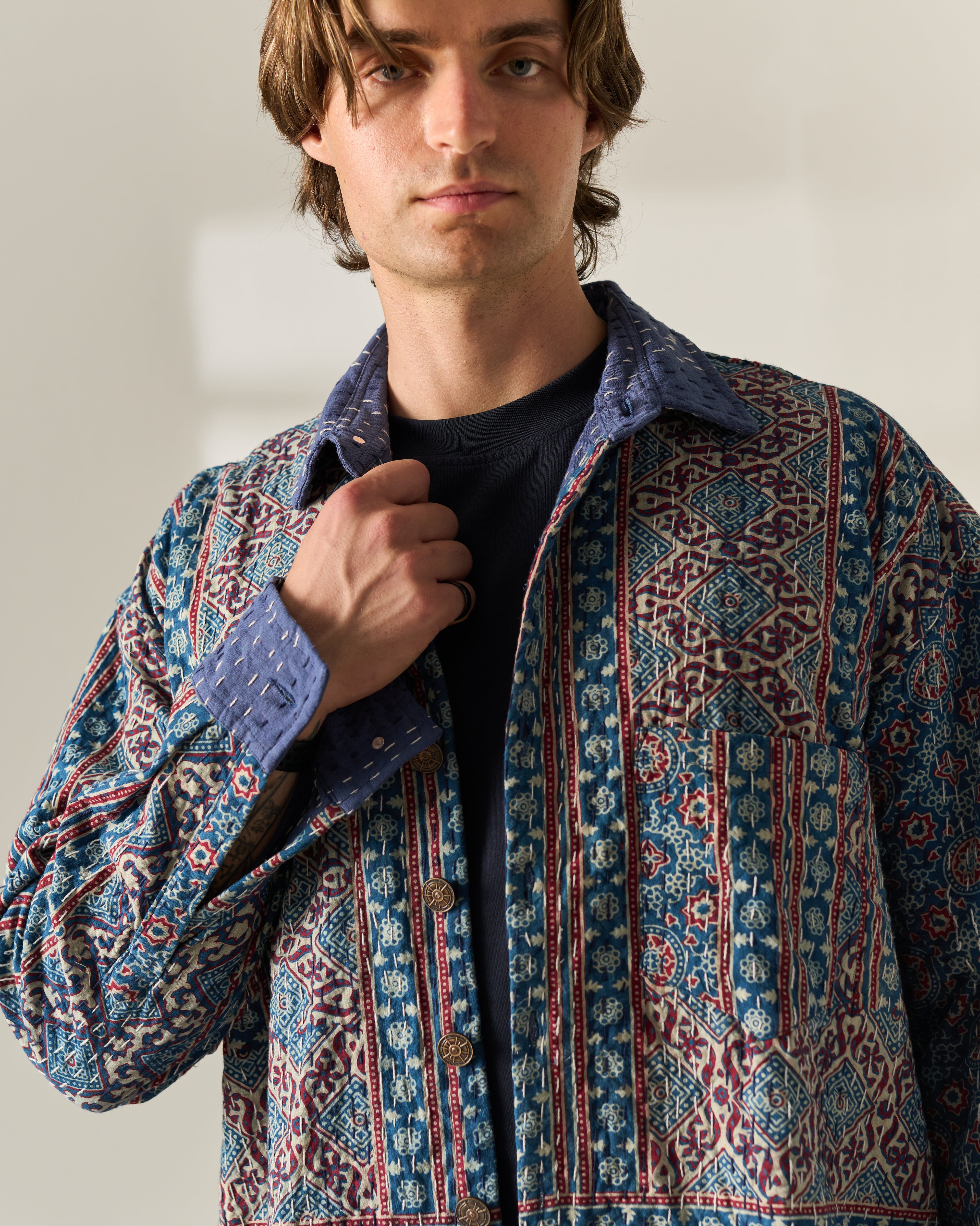 A man wearing a blue and red patterned button-up Hand Block painted Ajrakh kantha shirt jacket over a black t-shirt, photographed in a studio setting with a warm off-white background.