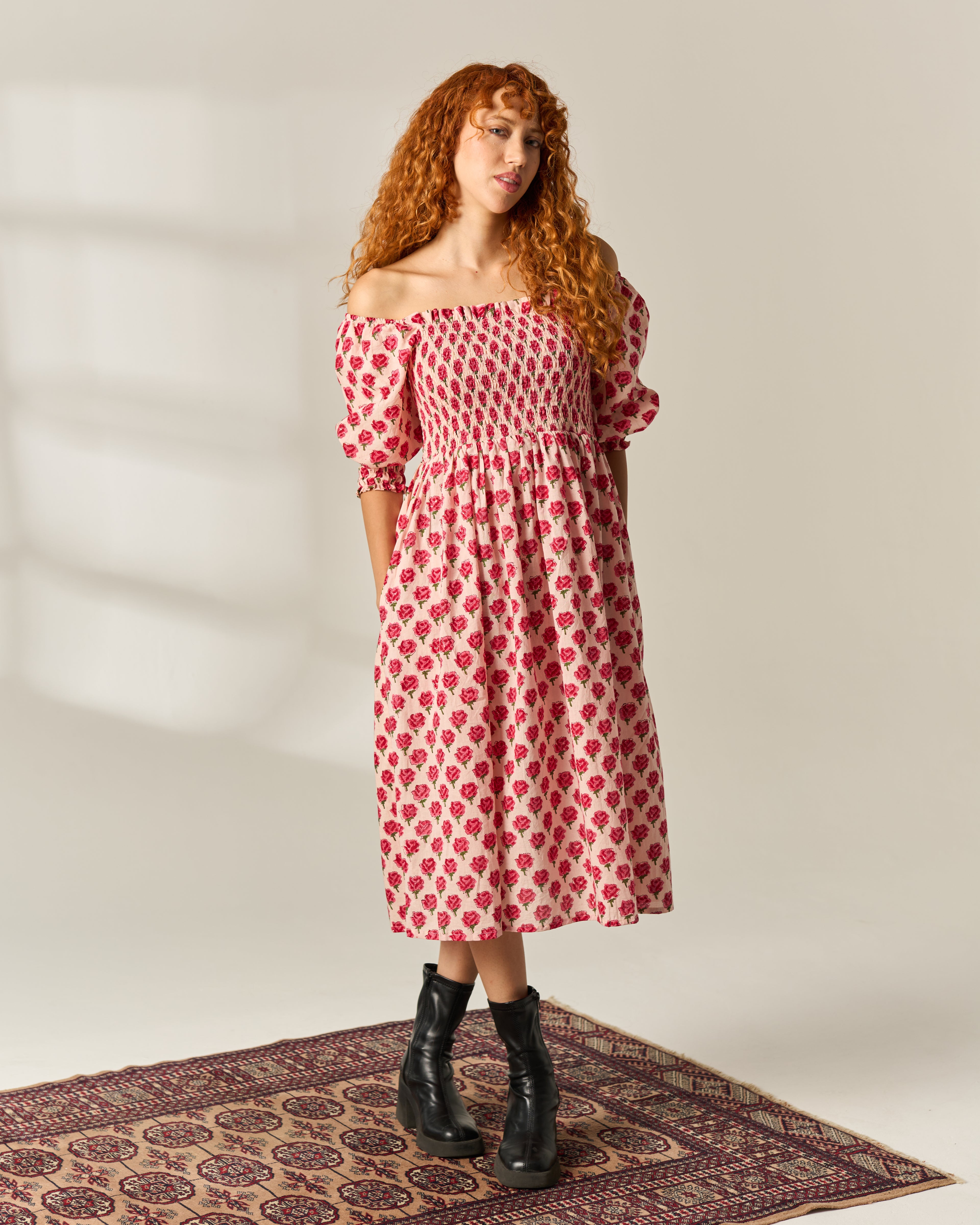 A woman with long curly red hair stands on a patterned rug wearing an off-the-shoulder midi dress with a red and white floral hand block print, puff sleeves, and black ankle boots against a plain light beige studio background.