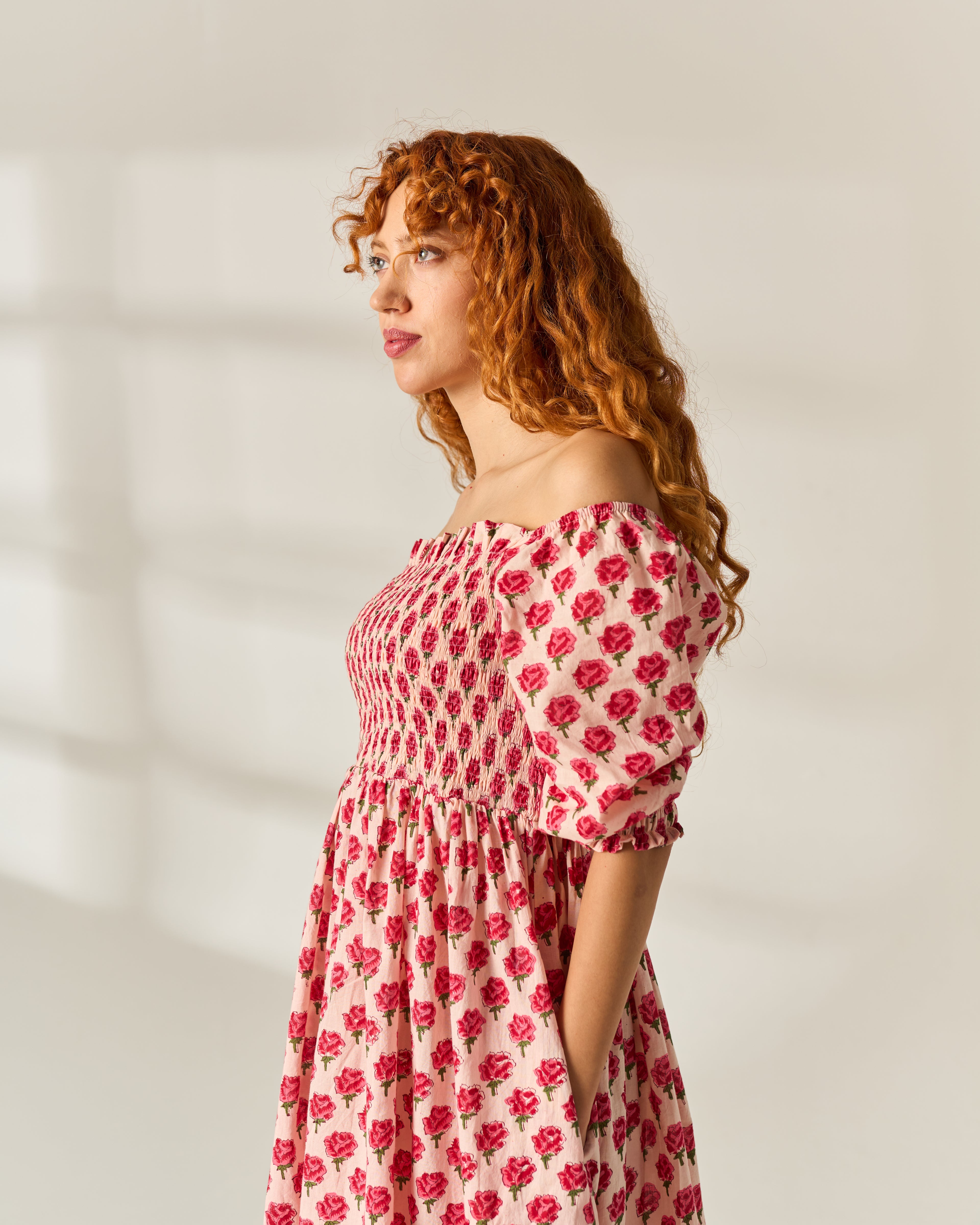 A woman with long curly red hair is shown in profile wearing an off-the-shoulder midi dress with a hand block pink and red floral pattern on a cream cotton base fabric, set against a warm off-white wall with soft window shadows.