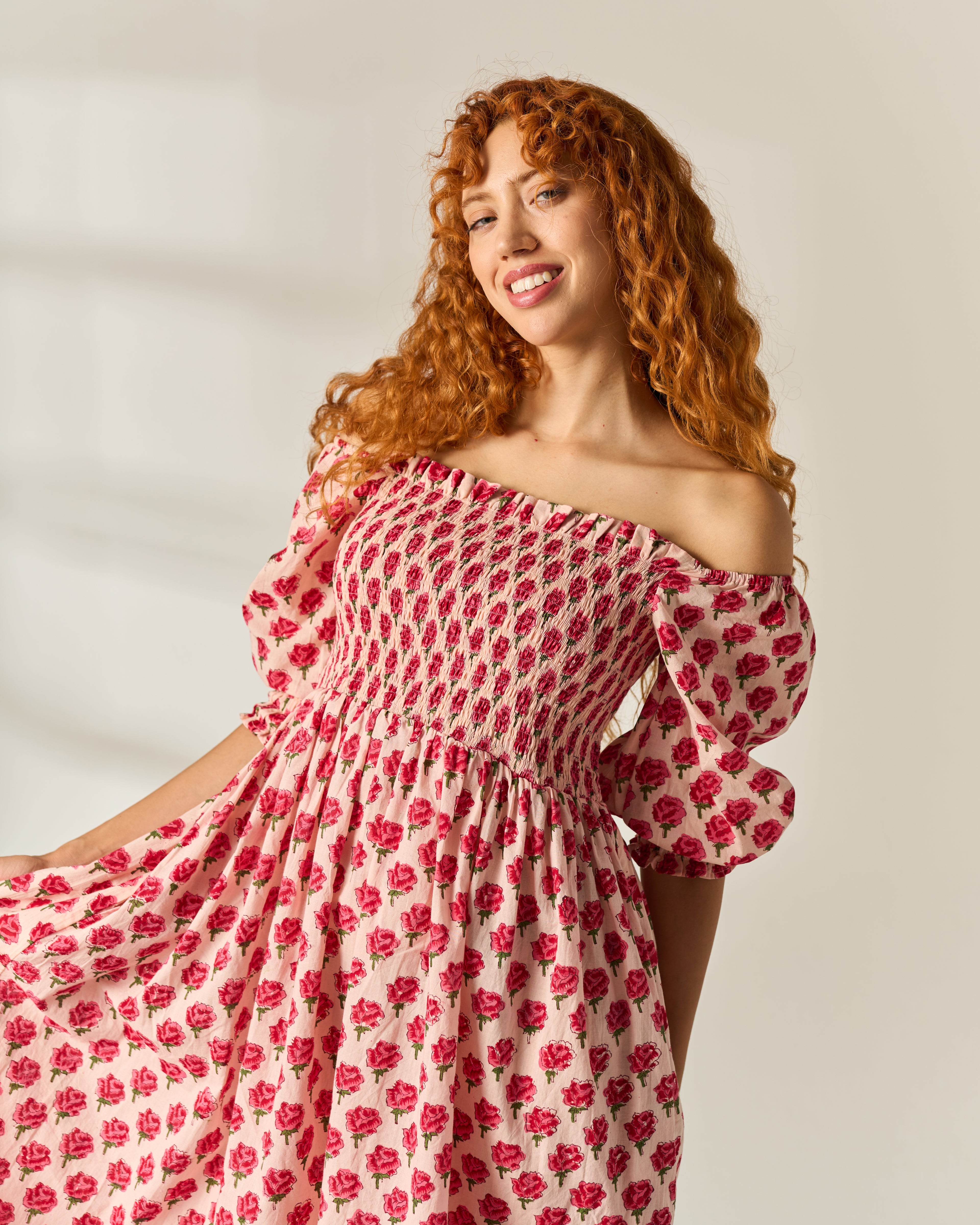 A smiling woman with curly red hair wearing an off-the-shoulder midi dress with a pink and red floral hand block print on a cream cotton base fabric, posed against a light neutral studio backdrop.