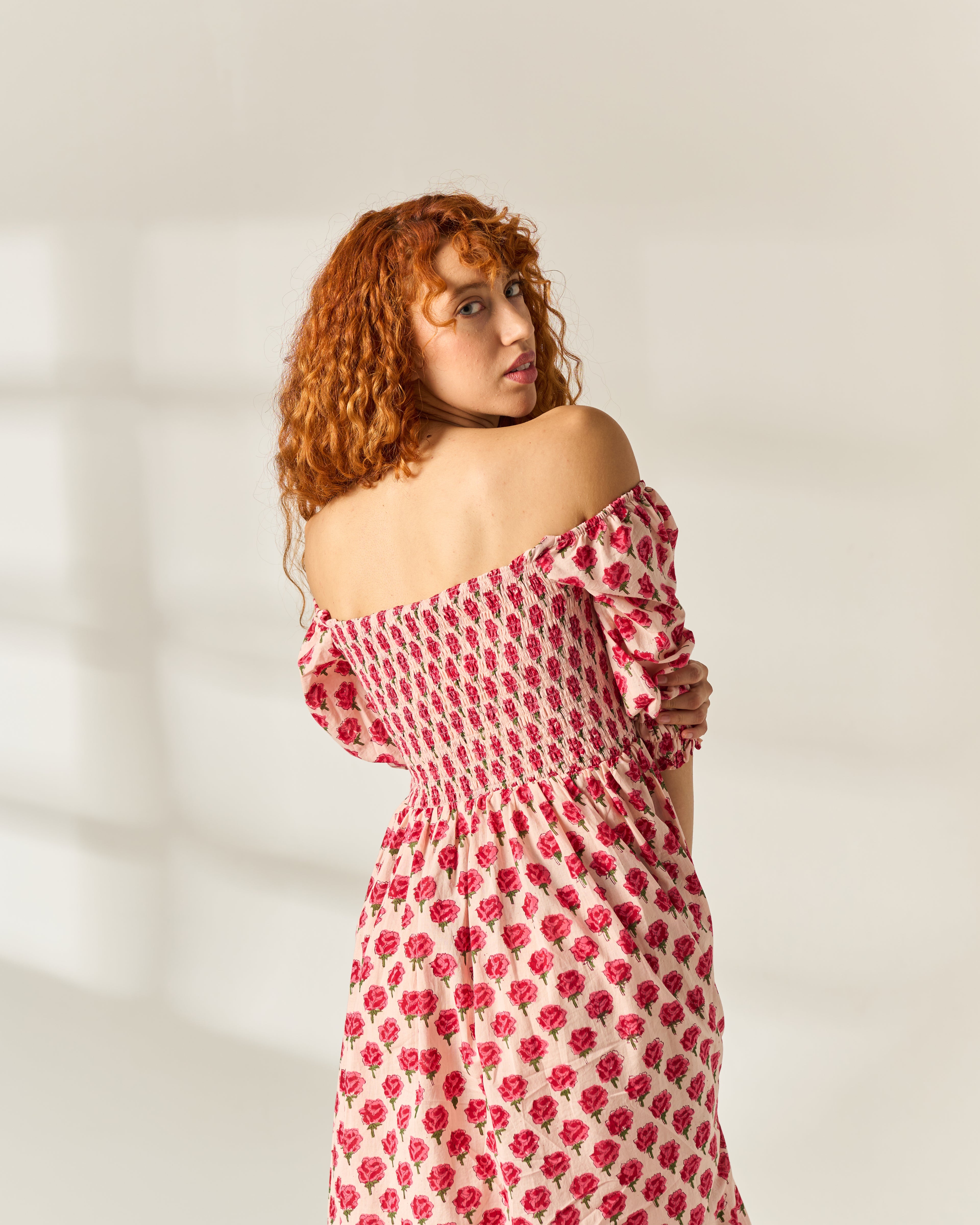 A woman with curly red hair wearing an off-the-shoulder midi dress with a red and white floral hand block print, photographed in a bright studio setting.
